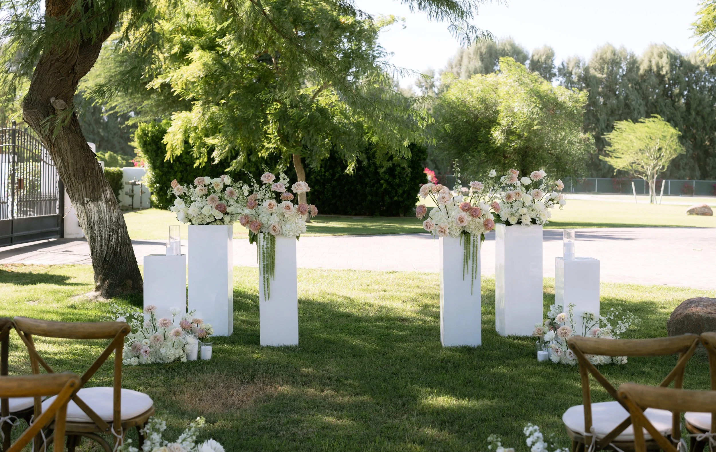 Outdoor wedding ceremony setup with white flower arrangements on tall white pedestals under a tree in a sunny garden.
