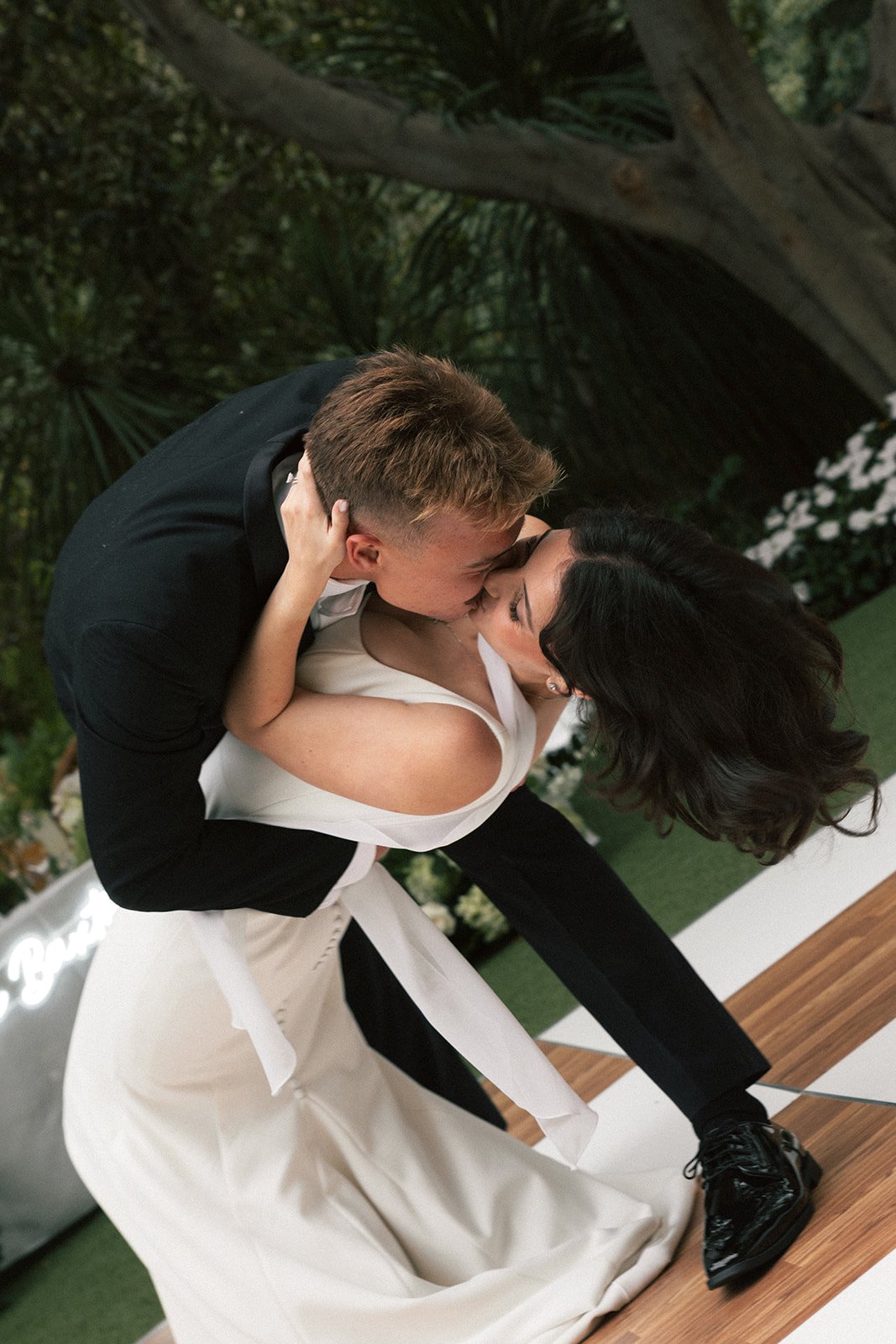 A couple in formal attire sharing a kiss on the dance floor, with the man in a black suit and the woman in a white dress in a decorated indoor setting.