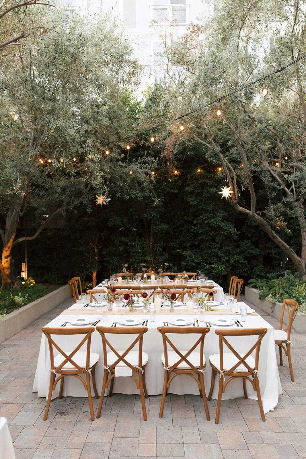 Elegant outdoor dining setup under string lights and star-shaped lanterns with a white tablecloth, floral centerpieces, and wooden chairs in a garden.