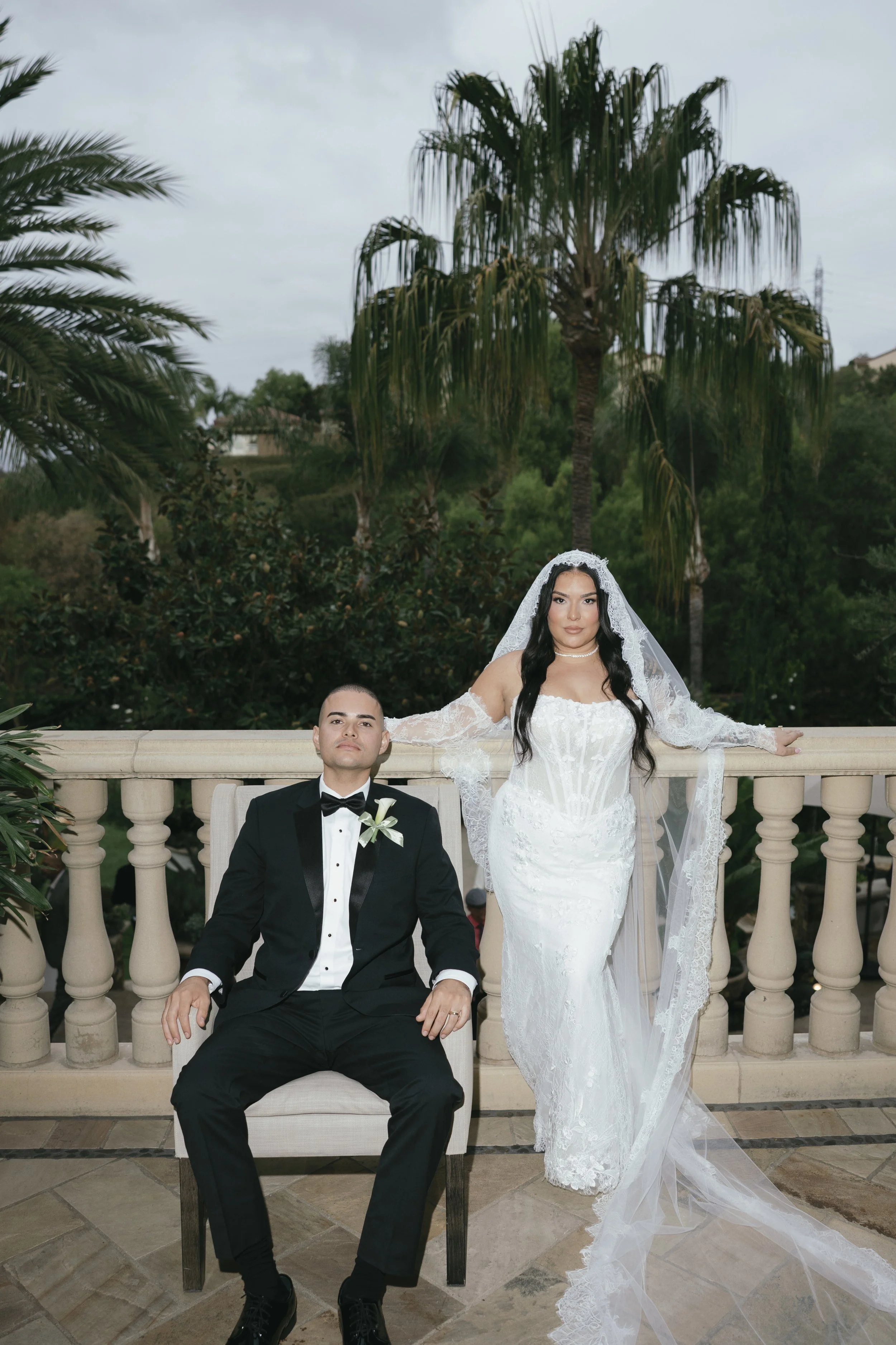 A bride in a white wedding gown with lace detailing and a veil standing behind a seated groom in a black tuxedo. The couple is outdoors on a terrace with a palm tree and lush greenery in the background.
