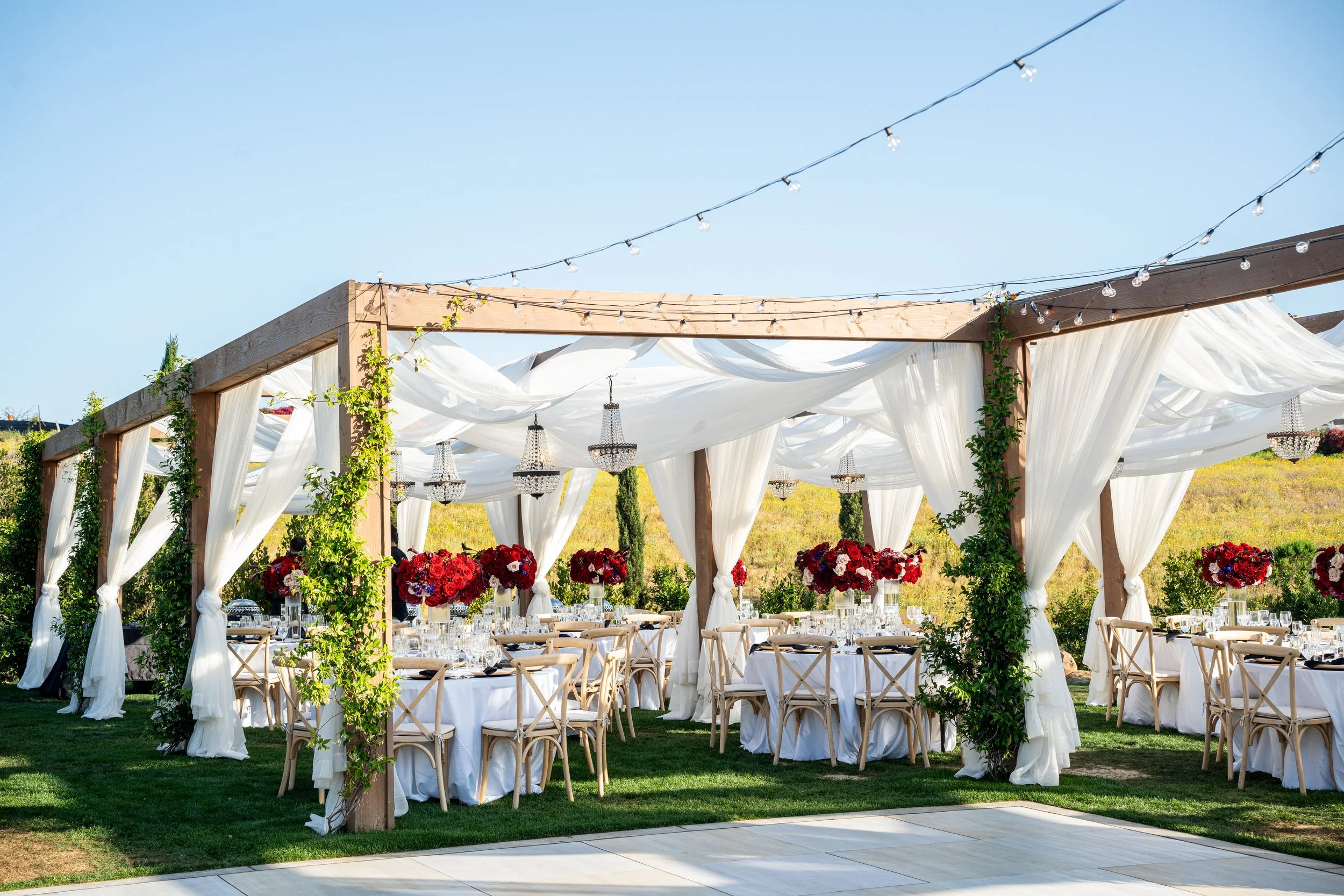 Outdoor wedding reception setup with round tables covered in white tablecloths, decorated with red floral centerpieces, under a wooden arch draped with white fabric, string lights, and chandeliers on a grassy area.