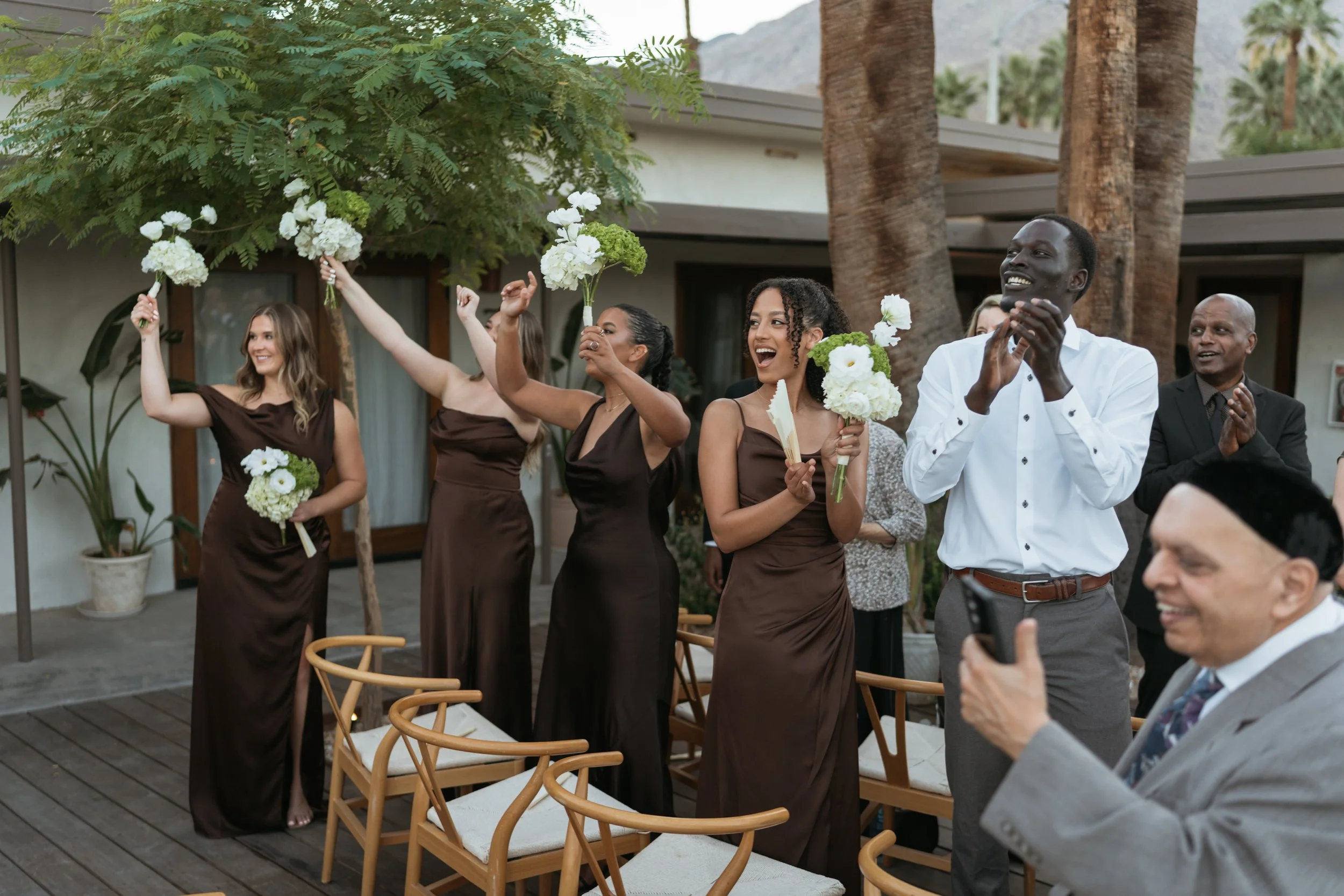 Group of women in matching brown dresses and men in white shirts and dark suits celebrating at an outdoor wedding or event, holding bouquets and clapping.