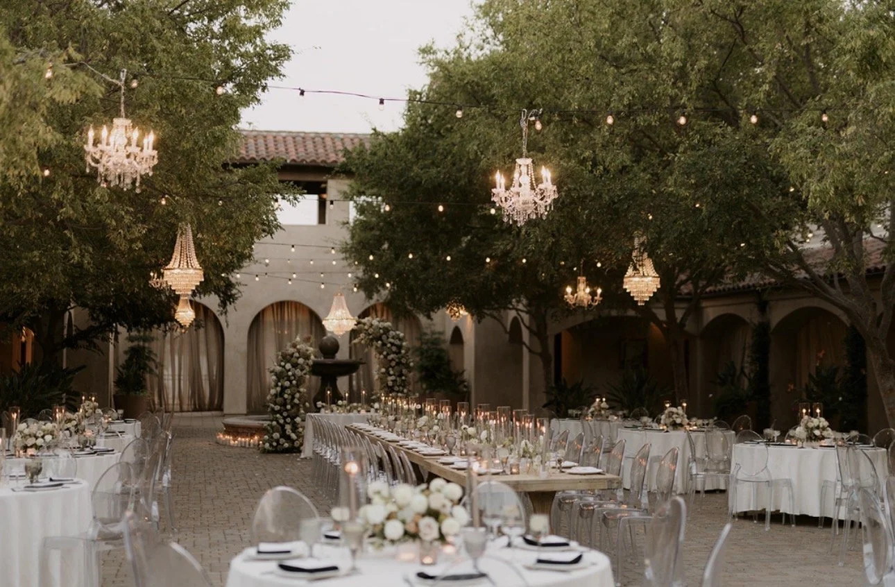 Elegant outdoor wedding reception area decorated with white flowers, candles, chandeliers, and string lights, surrounded by trees and a building with arched doorways.