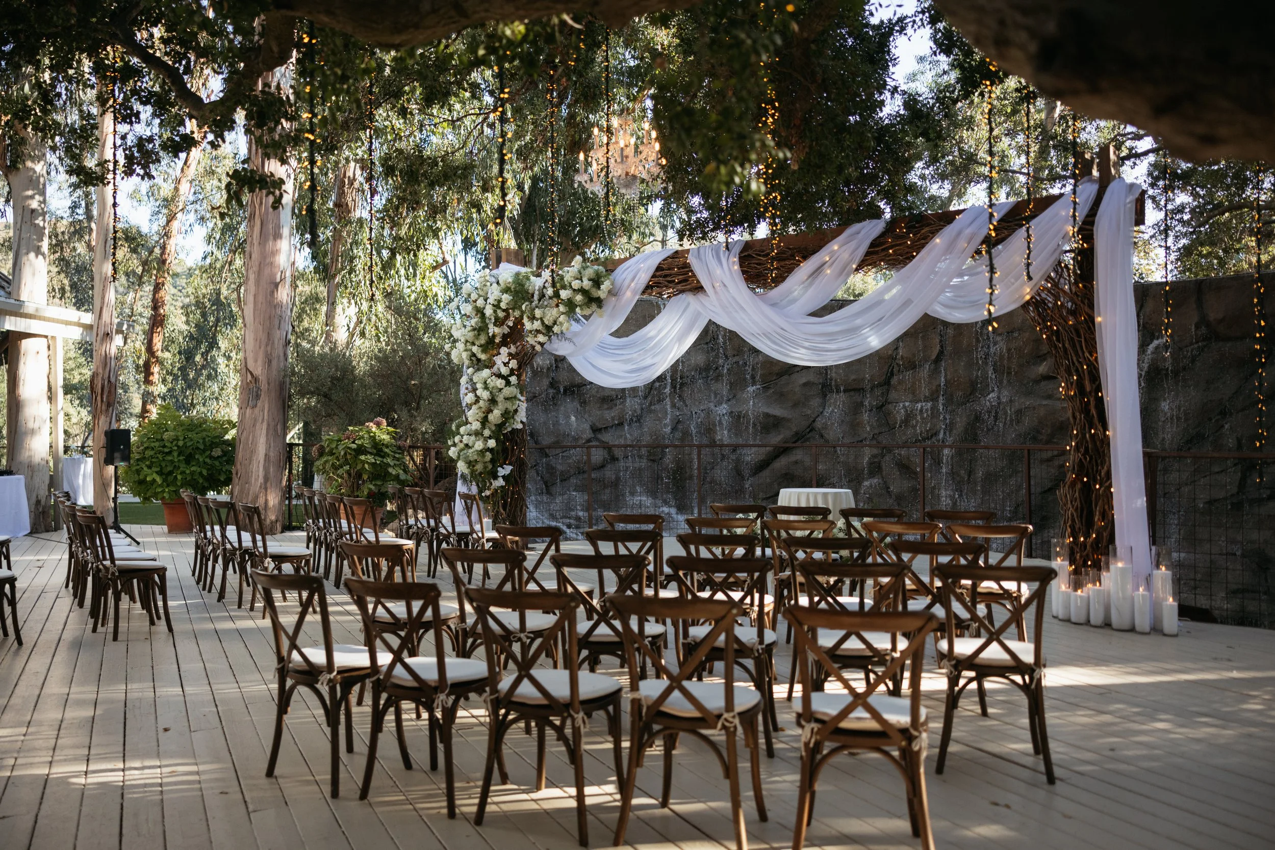 Outdoor wedding ceremony setup with rows of wooden chairs, floral arrangements, white drapes, string lights, and candles on the ground in a wooded area.