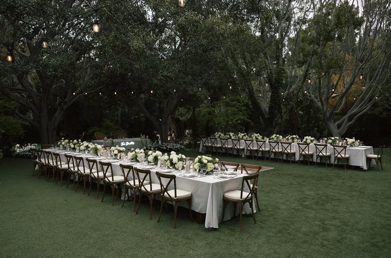 Outdoor wedding reception area with long dining tables decorated with white flower centerpieces, surrounded by wooden chairs, under string lights in a garden setting with large trees.