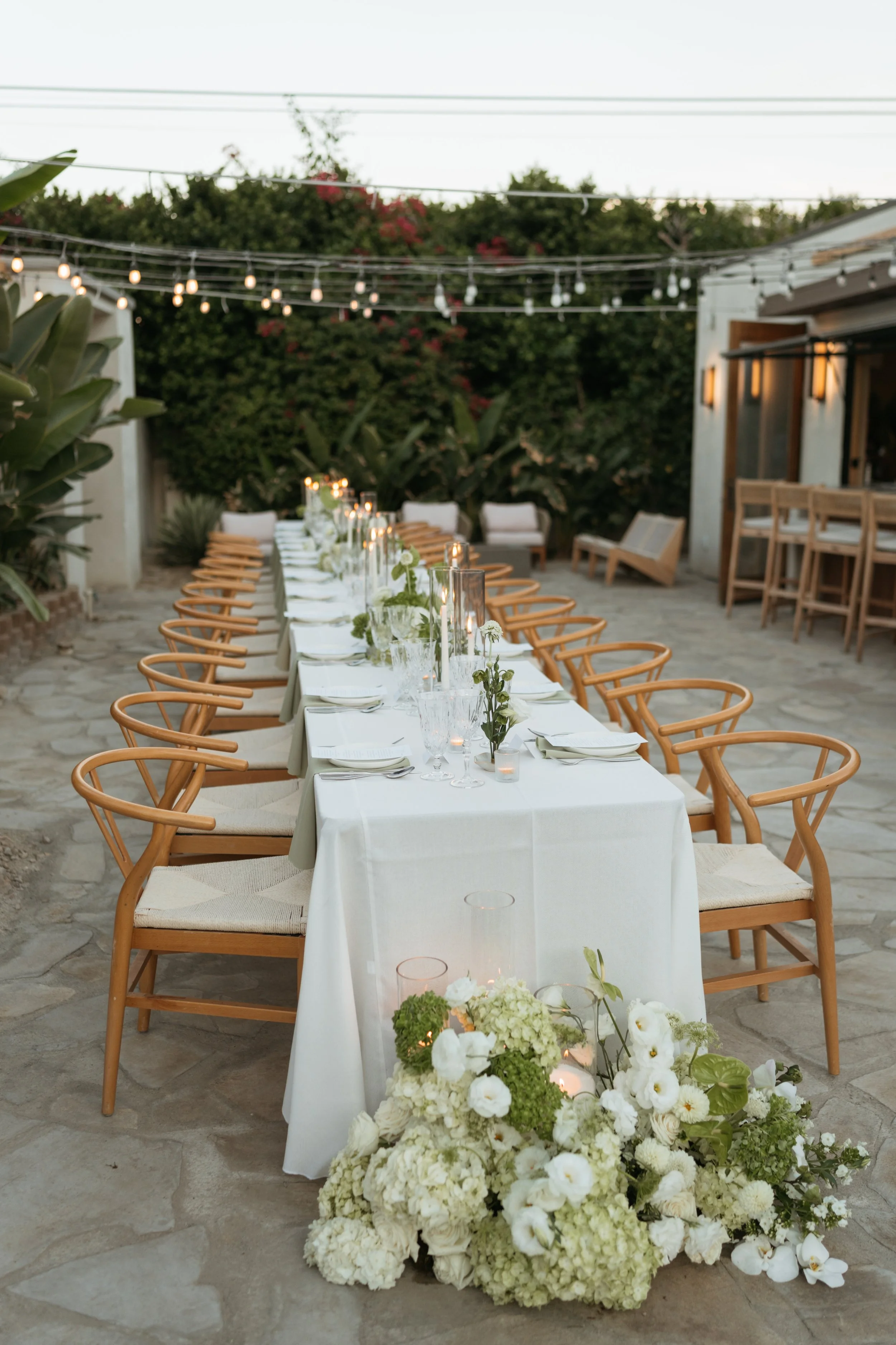 Decorated outdoor dinner table with white tablecloth, white floral arrangements, candle lanterns, and surrounding wooden chairs, set for an evening event.
