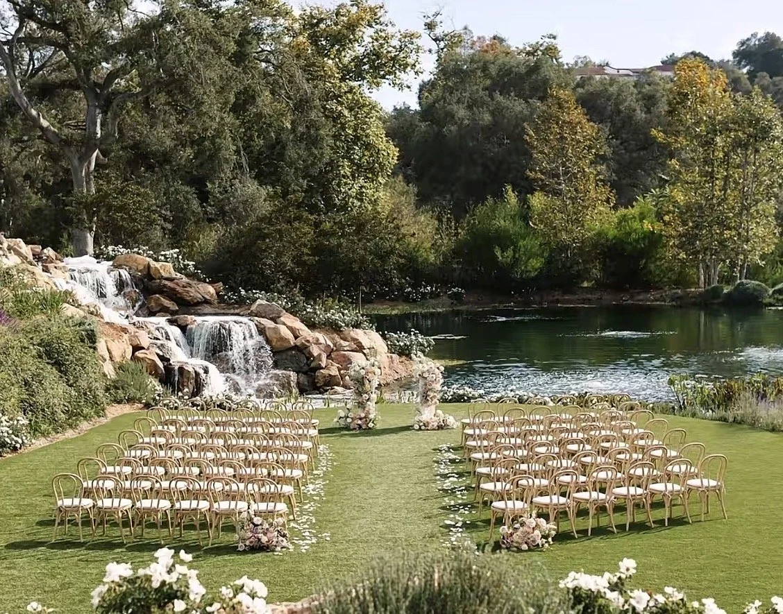 Outdoor wedding ceremony setup with chairs arranged on a grassy area near a lake, with a waterfall, trees, and floral decorations.