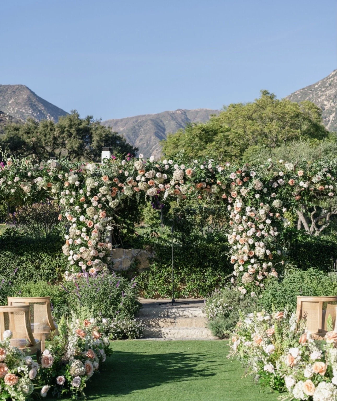 A floral wedding arch decorated with white and pink flowers in an outdoor garden with mountains in the background.