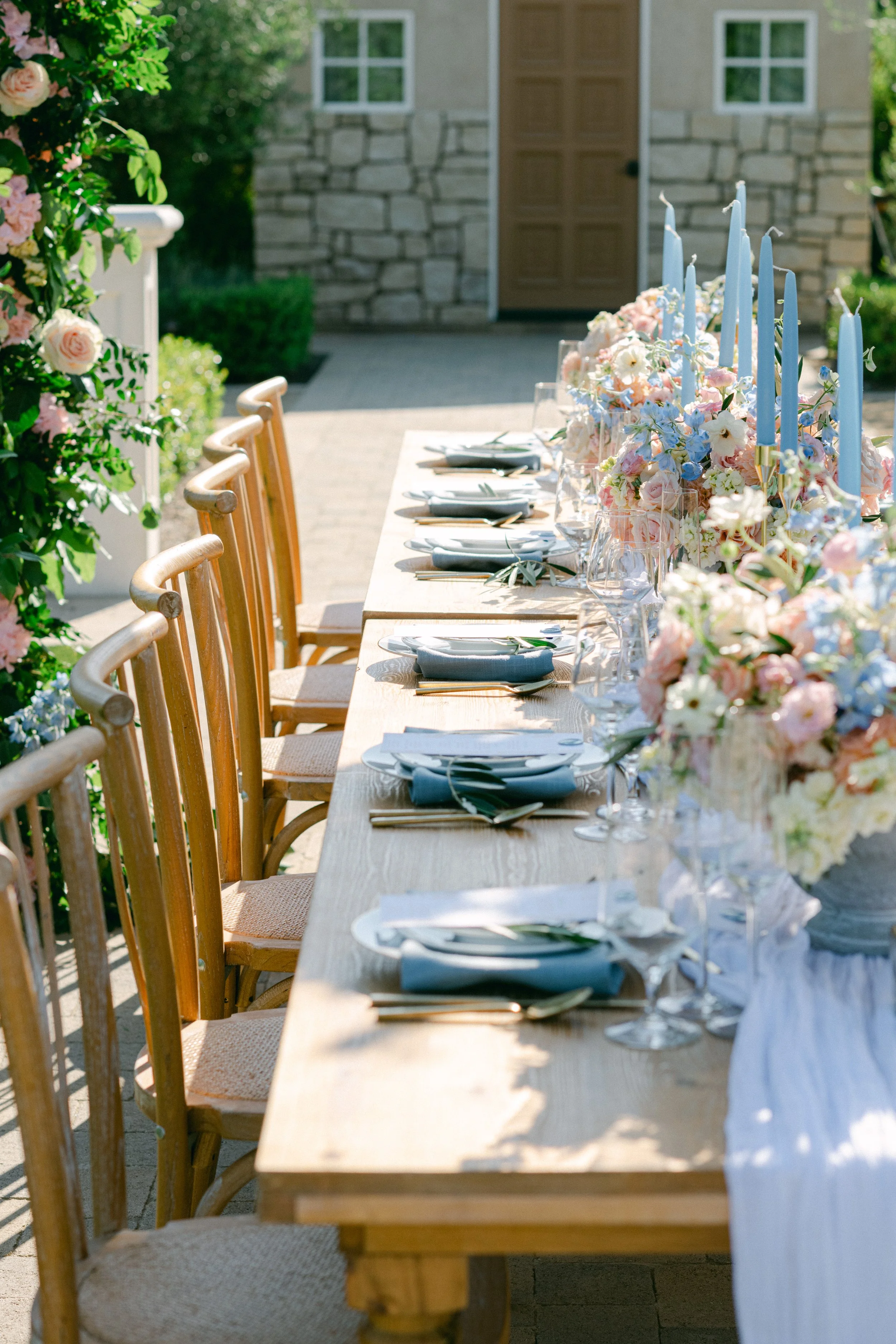 A long outdoor dining table set with plates, silverware, wine glasses, and blue napkins, decorated with pastel flower arrangements and tall blue candles, on a sunny day.