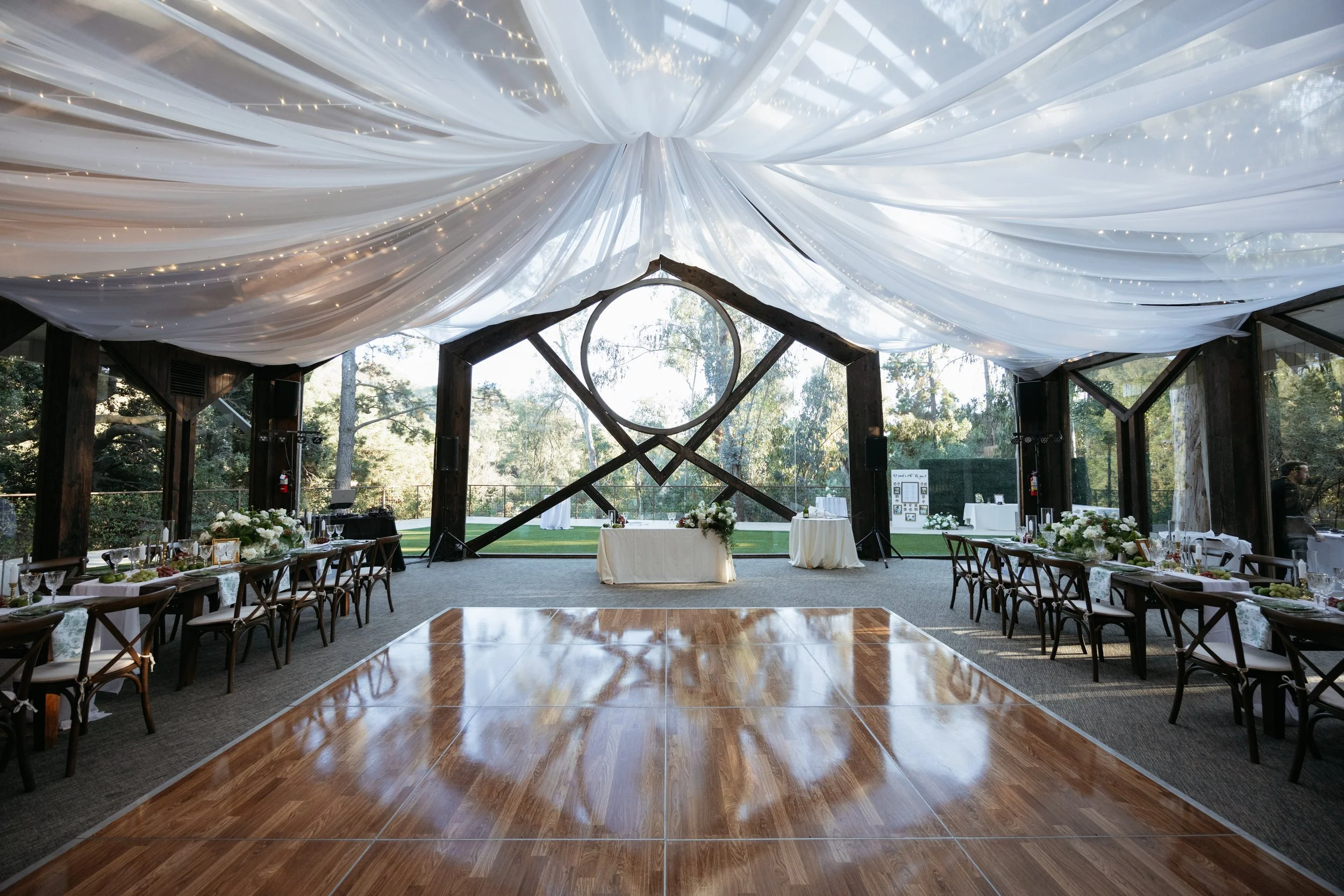 Wedding reception area with decorated tables, floral centerpieces, a dance floor, and a transparent ceiling with draped fabric and string lights, overlooking a garden outside.