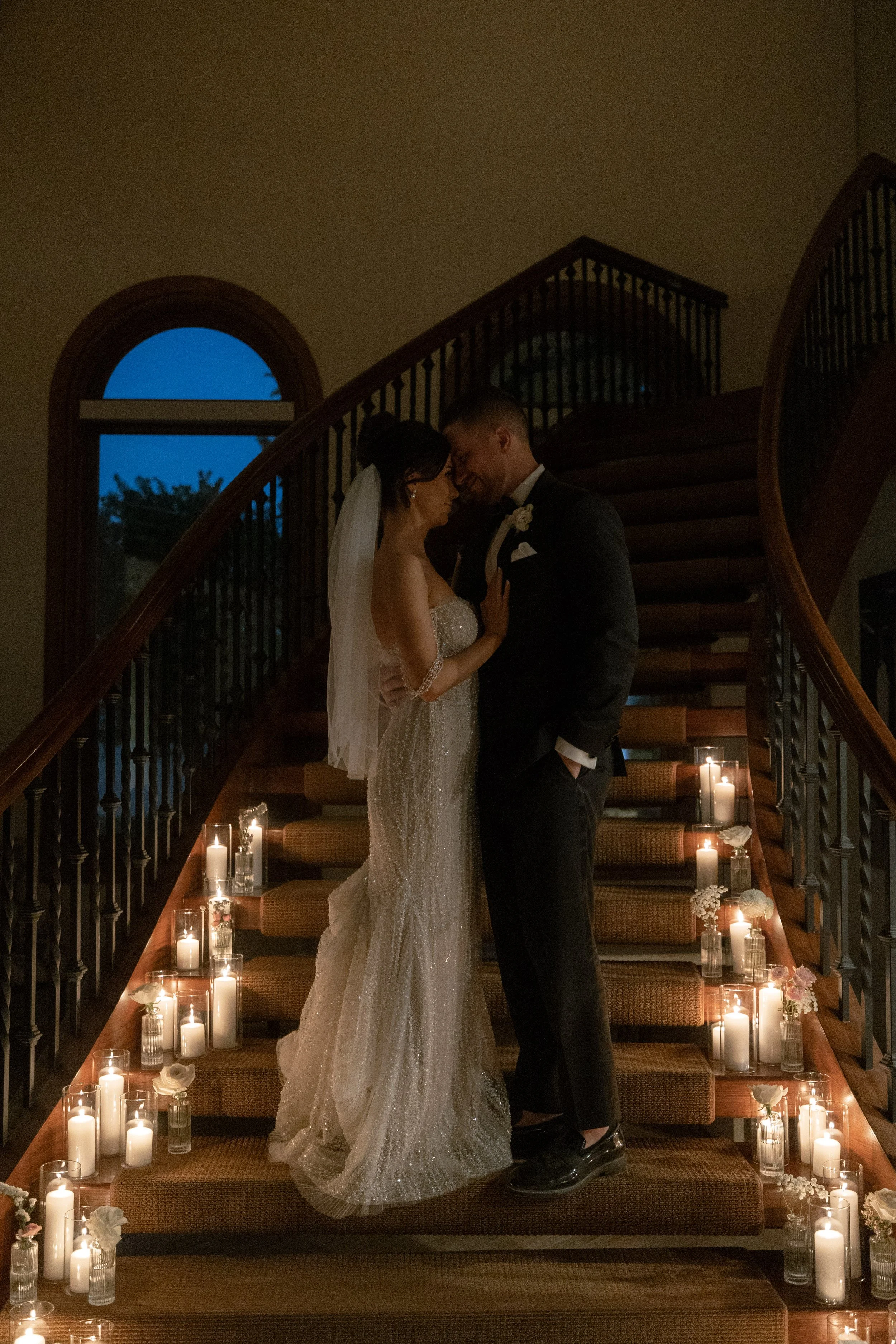 A bride and groom standing close together on a staircase adorned with candles and flowers during a wedding at night.