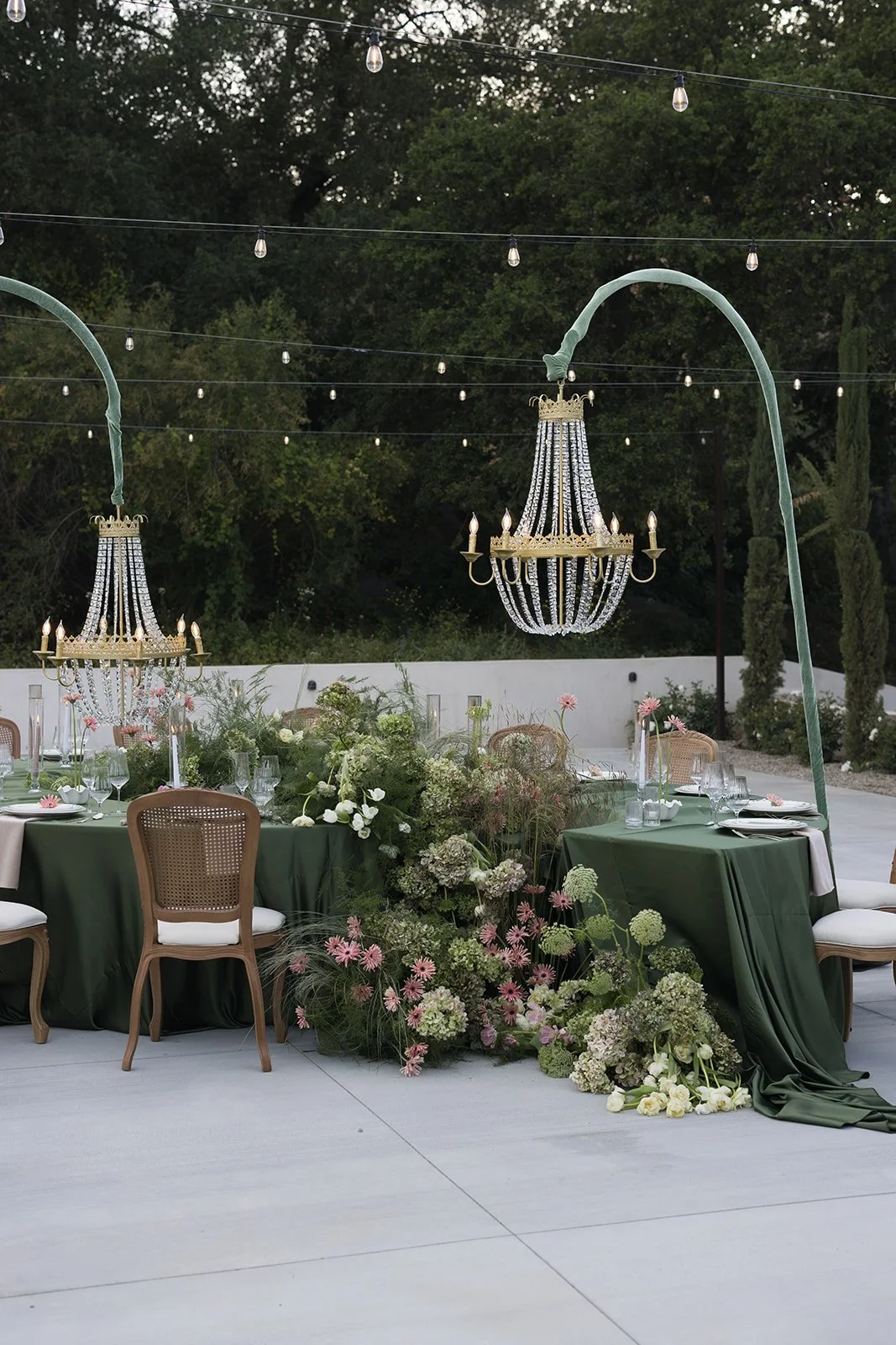 Outdoor event setup with round tables covered in green tablecloths, decorated with floral centerpieces, and illuminated by vintage chandeliers hanging from green arches. String lights are overhead, and a white wall and trees are in the background.