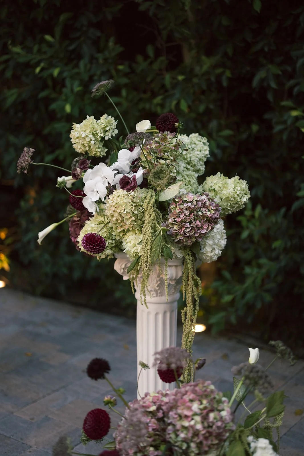 A tall white pedestal holding a large arrangement of white, green, and burgundy flowers, including hydrangeas, orchids, and other blooms, set outdoors on a stone patio with a green leafy background.