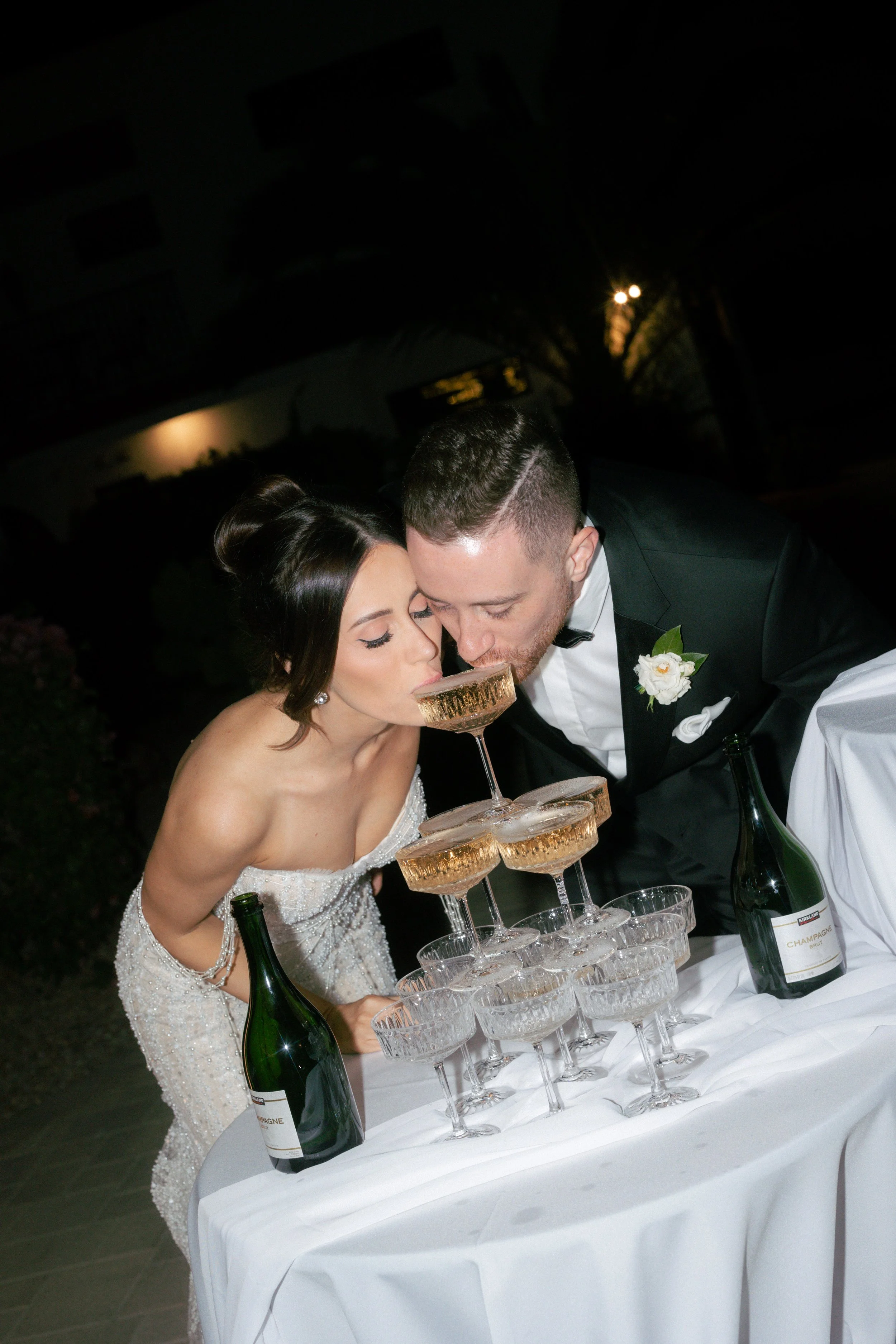 Bride and groom in formal attire pour champagne into a tower of glasses at their wedding celebration.