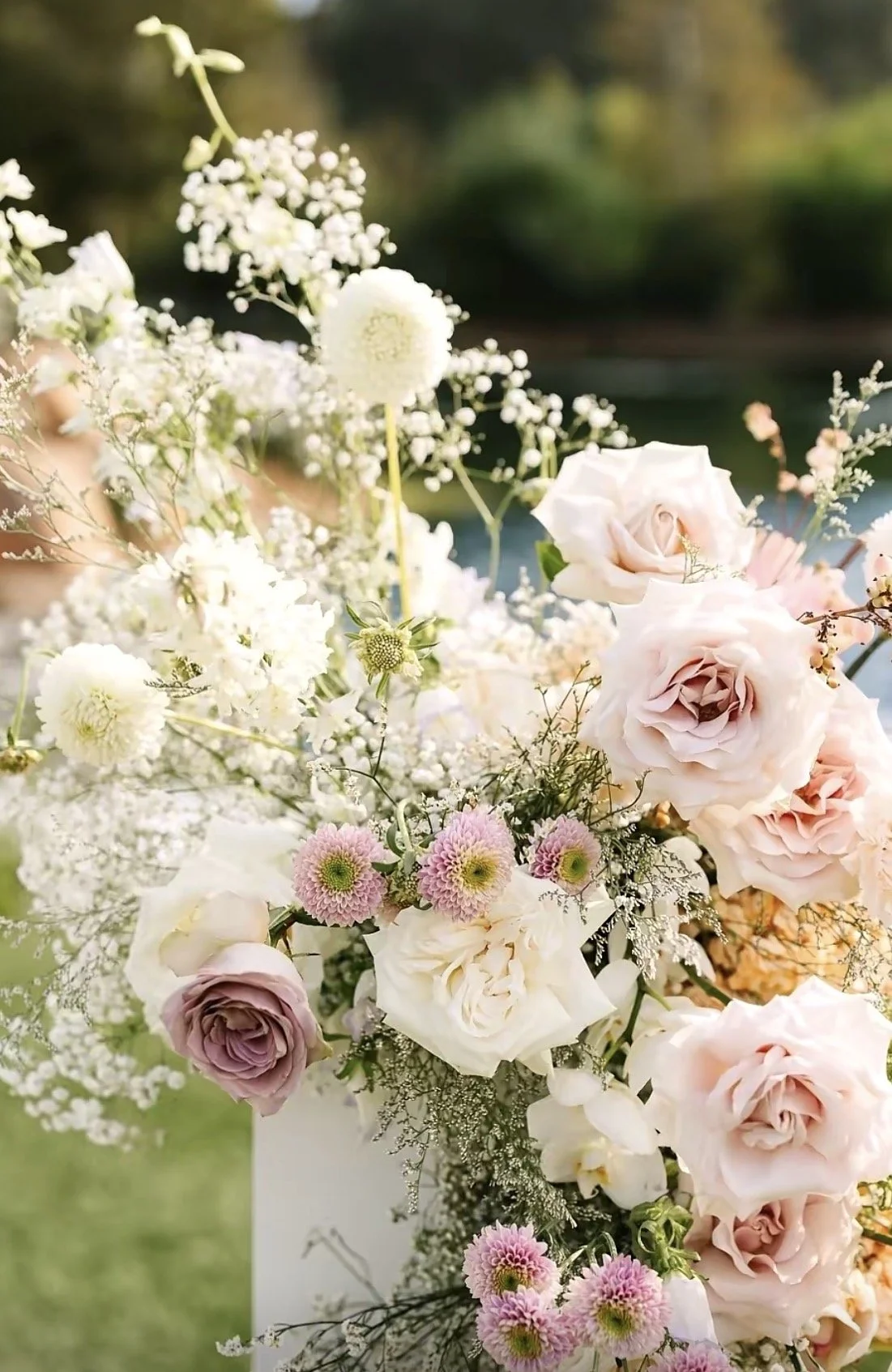 A floral arrangement with light pink roses, white roses, and pink and white chrysanthemums, accented with small white filler flowers and greenery, set outdoors.