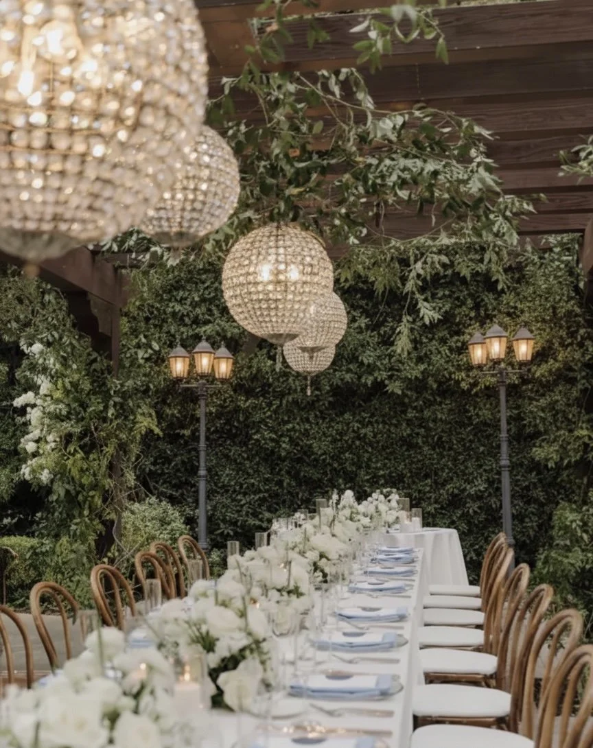 Long banquet table set for a formal event, with white tablecloths, white floral centerpieces, and place settings. Overhead, there are woven chandelier lights and hanging greenery. The background features lush green foliage and vintage-style lampposts.
