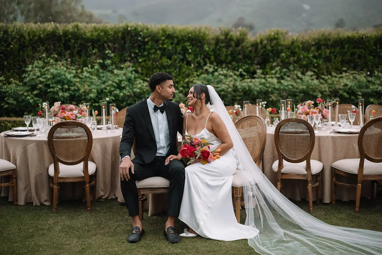 A couple in wedding attire, sitting close to each other outdoors at a decorated reception table. The bride holds a bouquet of flowers and smiles at the groom. The reception table is adorned with floral centerpieces, candles, and glassware, amidst lus