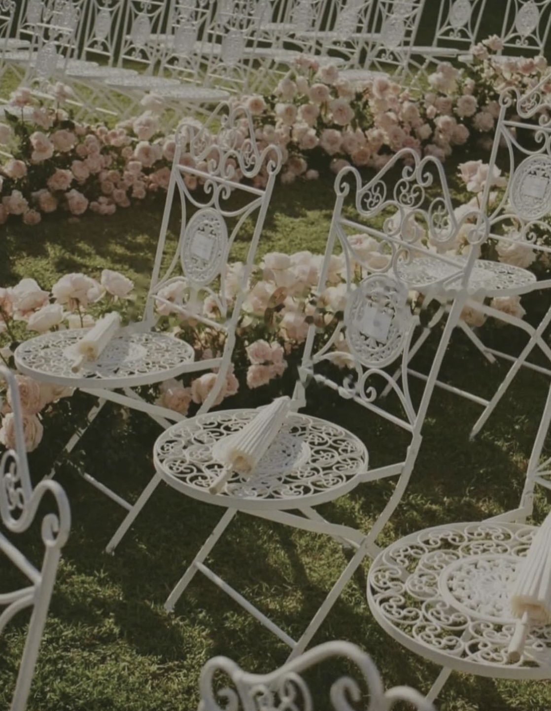 White ornate metal chairs with rolled paper scrolls on their seats, set outdoors on grass with pink roses and flowers arranged in the background.