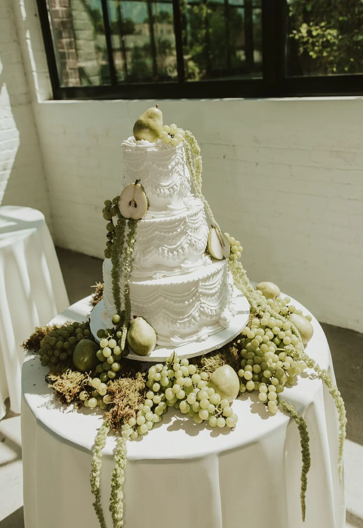 A white wedding cake decorated with white and green fruits and draped in white icing, surrounded by grapevine-like decorations on a round table.