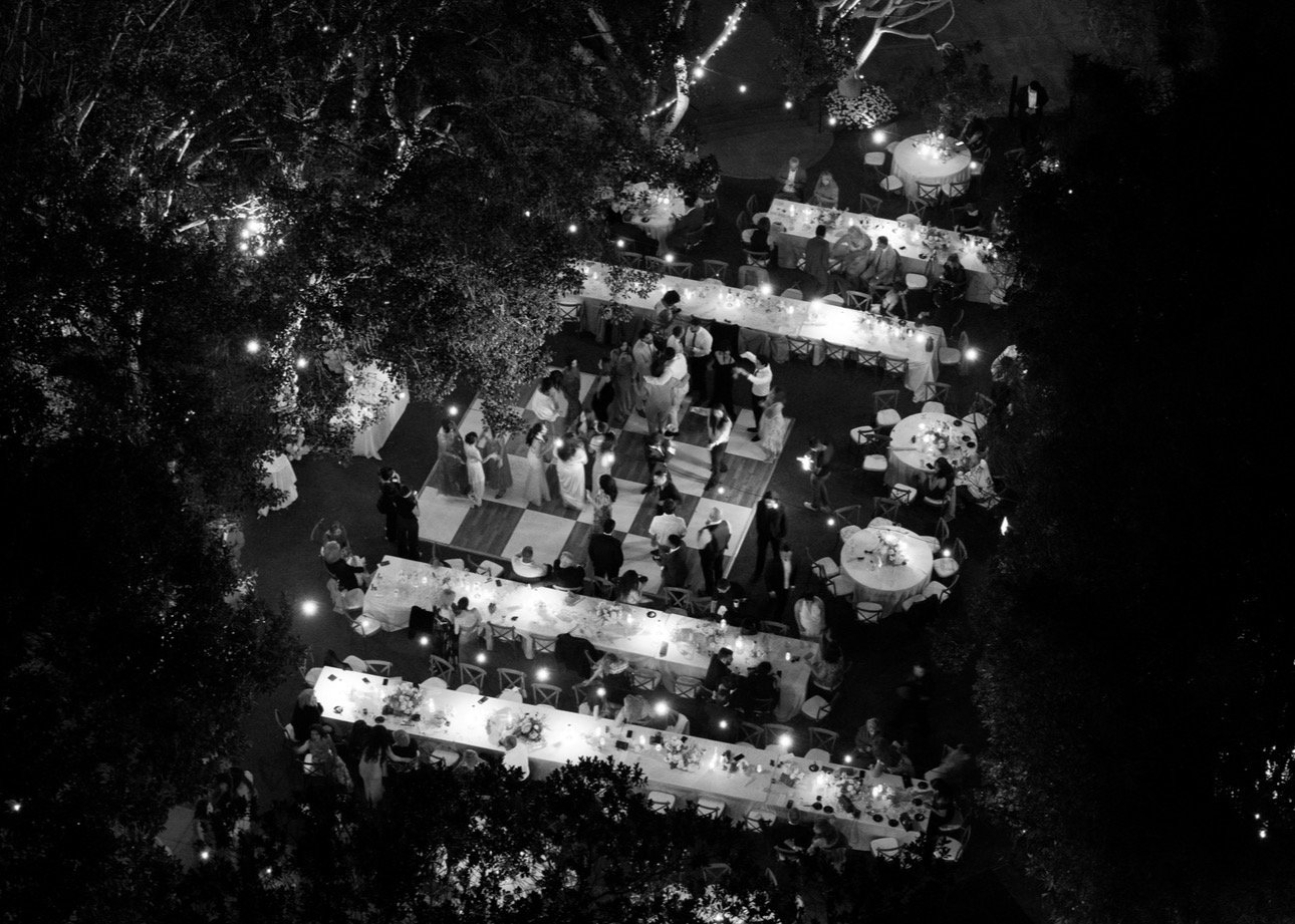 Dark aerial view of a nighttime outdoor wedding reception with illuminated tables and a dance floor surrounded by trees and guests