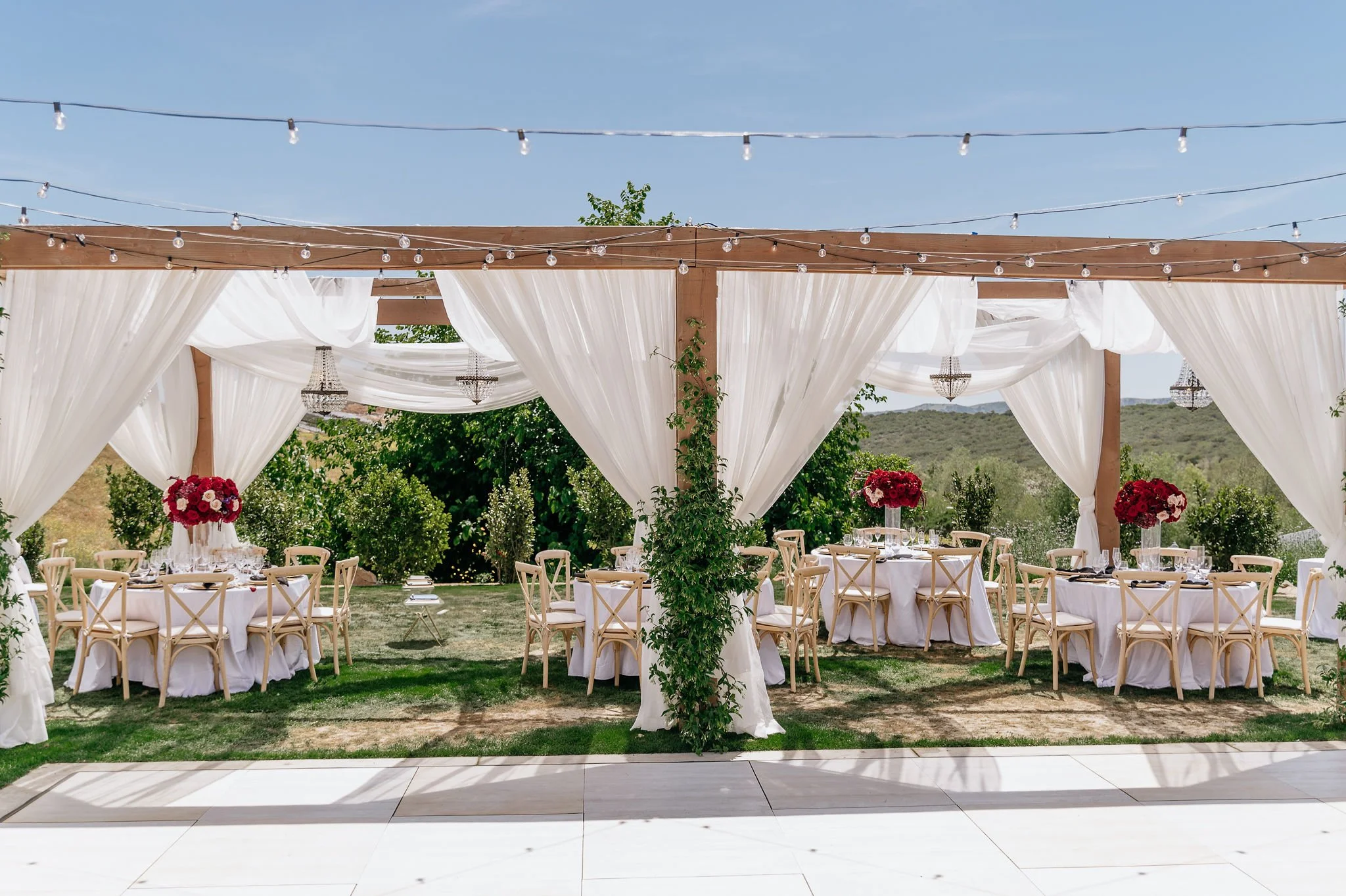 Outdoor wedding reception setup with round tables, white tablecloths, pink and red floral centerpieces, string lights, chandeliers, and white draped curtains under a wooden canopy with greenery in background.