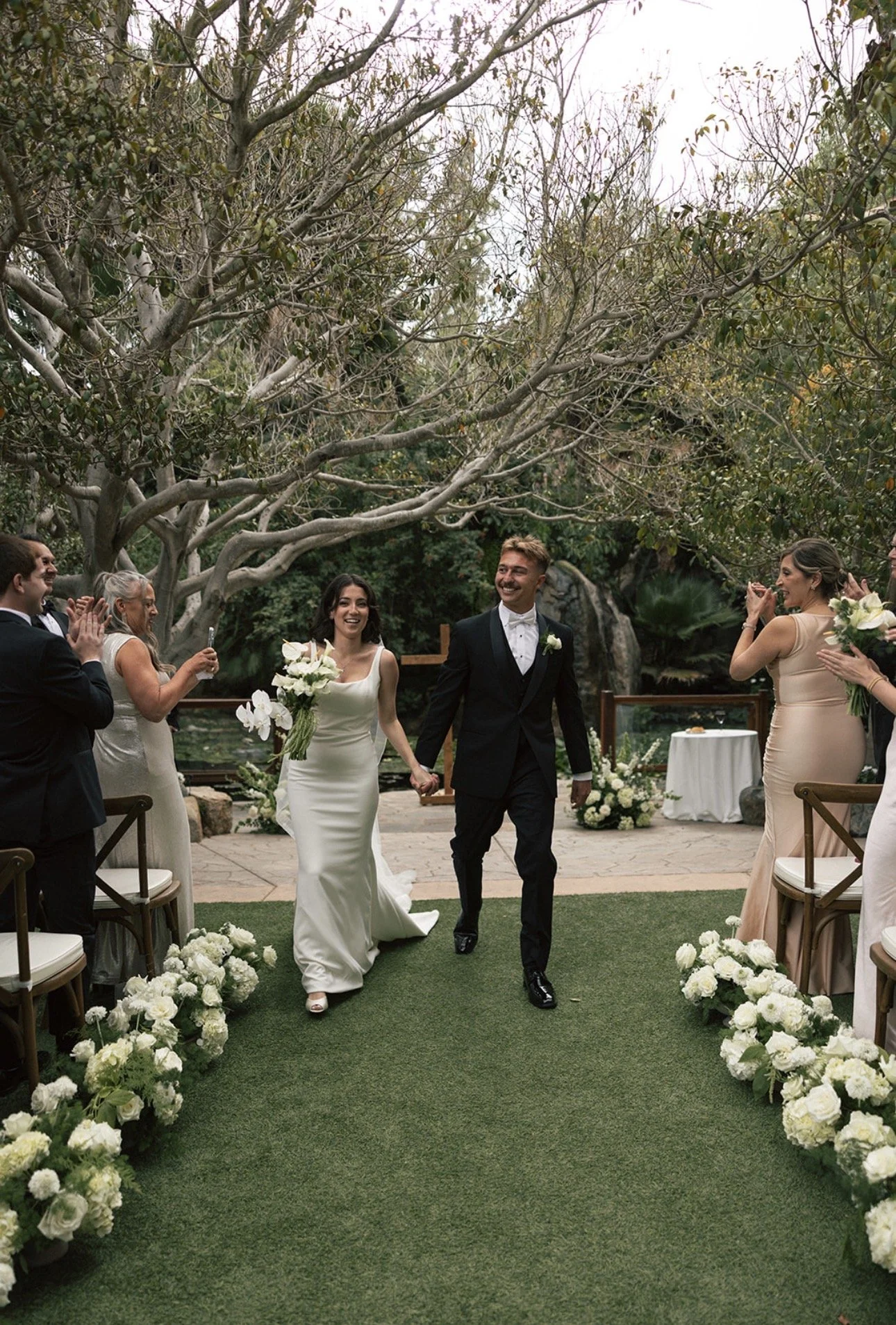 A newlywed couple holding hands and smiling as they walk down the aisle at their outdoor wedding ceremony, surrounded by friends and family.