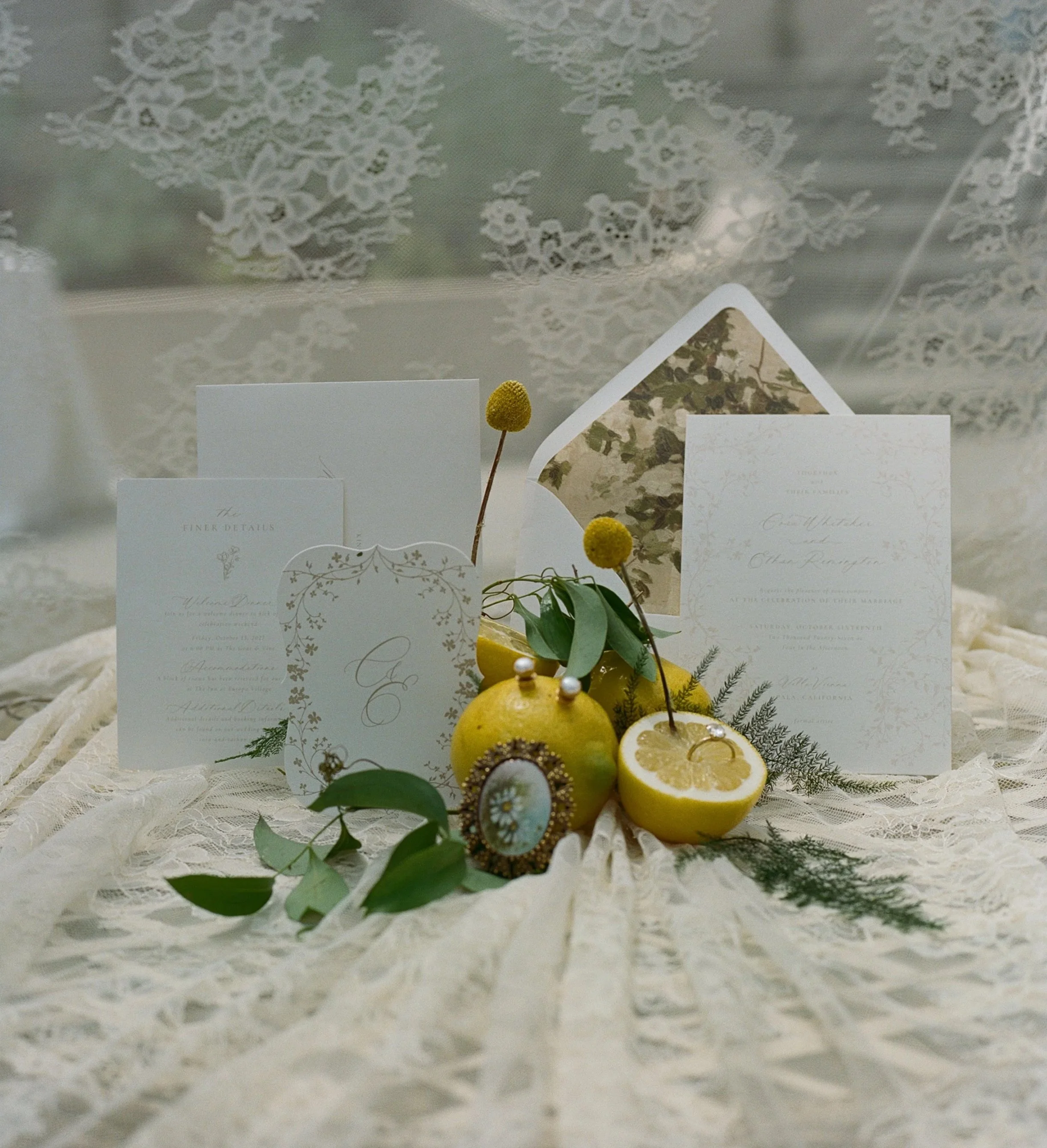 Elegant wedding invitation cards surrounded by greenery, lemons, and decorative jewelry on lace fabric with a lace ceiling.