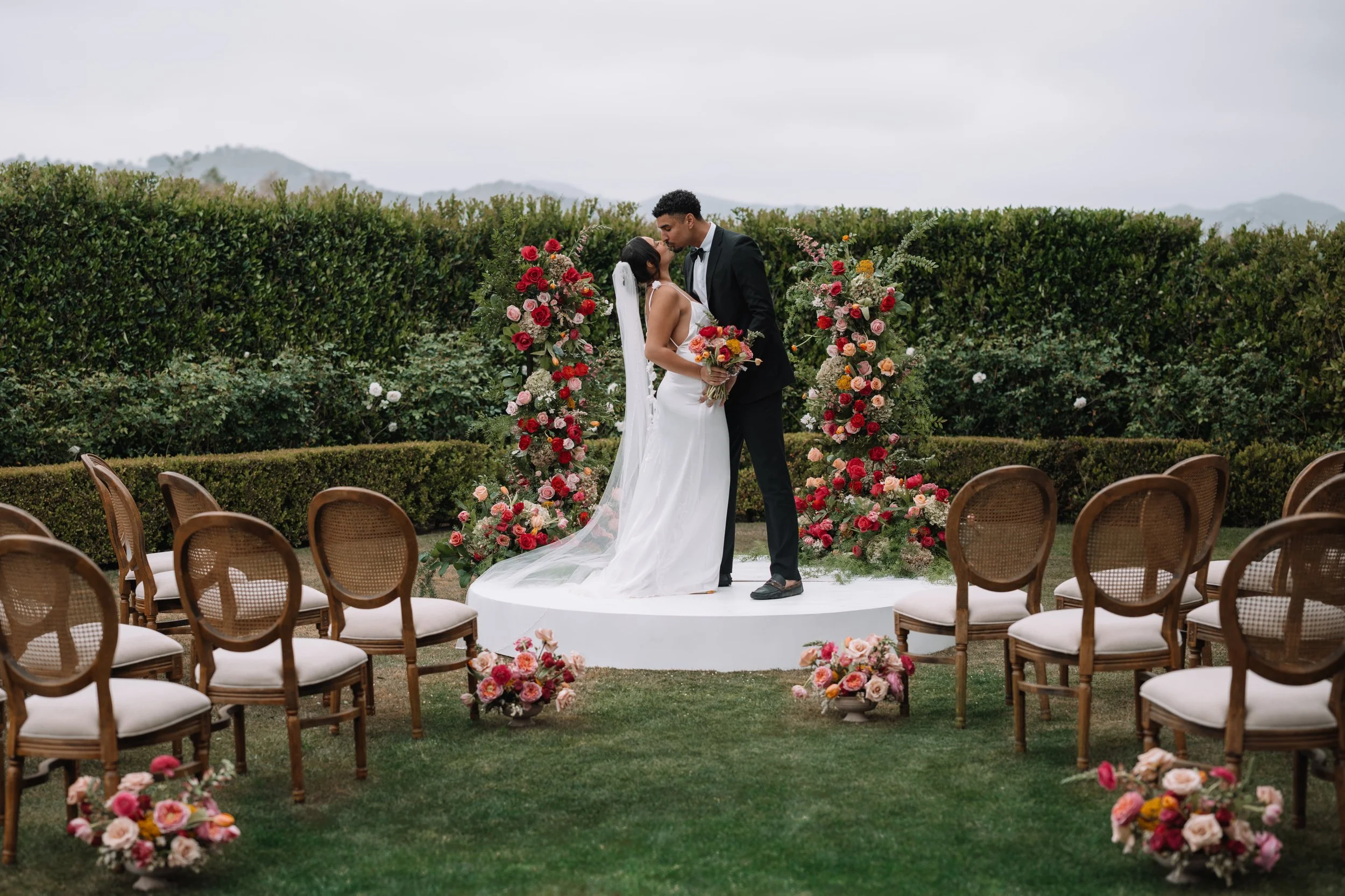 A couple at their outdoor wedding ceremony, kissing, with floral arrangements and chairs surrounding them on a grassy area.