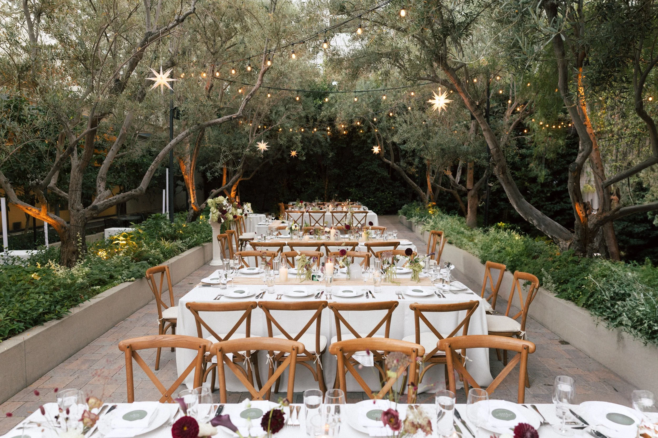 A beautifully decorated outdoor dining setup in a garden, with long white tablecloth-covered tables, wooden chairs, floral centerpieces, and string lights hanging among trees.