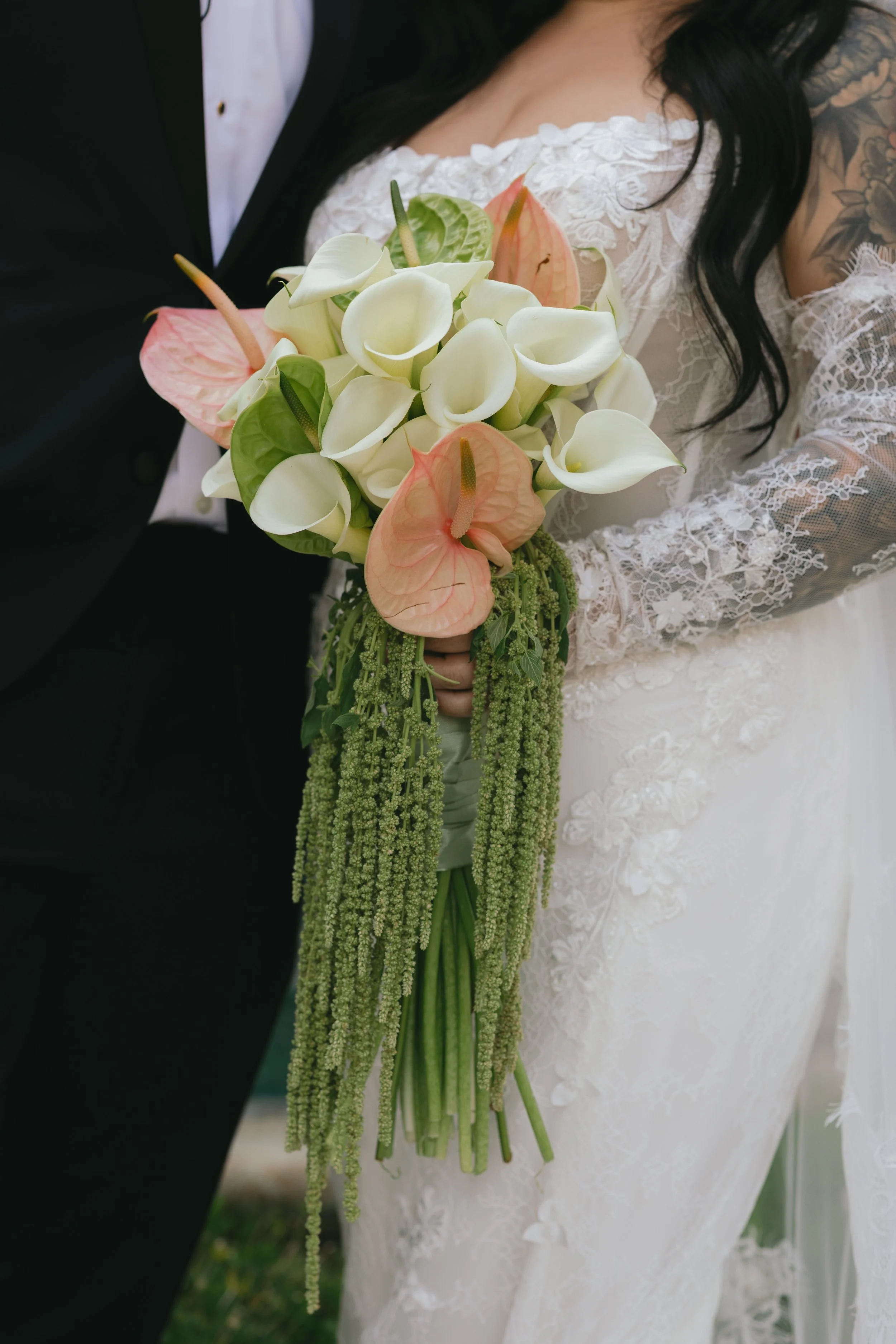 Close-up of a bride in lace wedding dress holding a cascading bouquet of white calla lilies, pink anthuriums, and greenery, standing next to a groom in a black tuxedo.