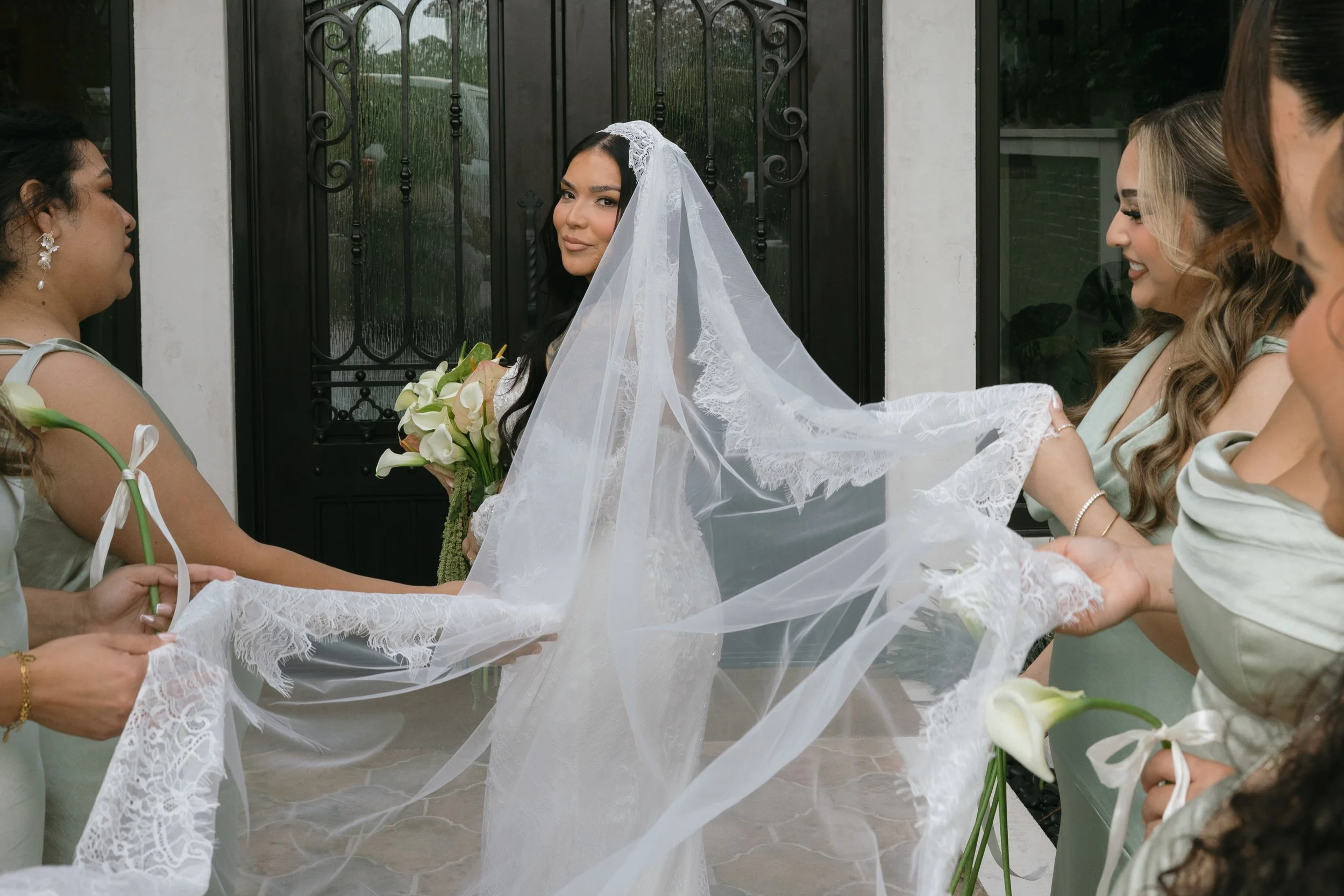 Bride holding a bouquet of white calla lilies and friends adjusting her wedding veil outside a door.