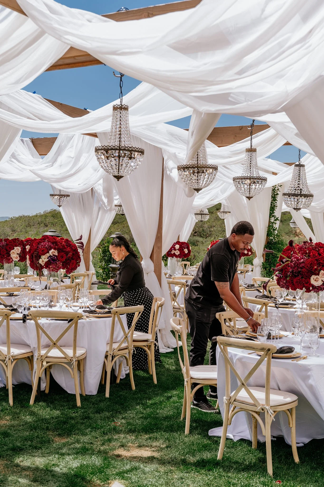 Event setup outdoors with tables, white tablecloths, and large red floral centerpieces, under white draped fabric and chandeliers, with people preparing the tables against a green landscape and blue sky.