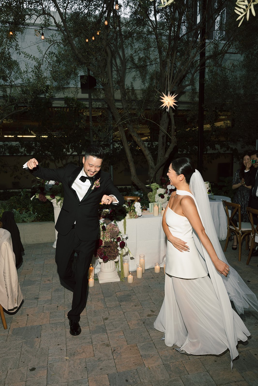 A joyful wedding celebration with a bride in a white gown and veil and a groom in a black tuxedo dancing happily indoors at night, surrounded by floral arrangements and candles, with a decorated outdoor setting in the background.
