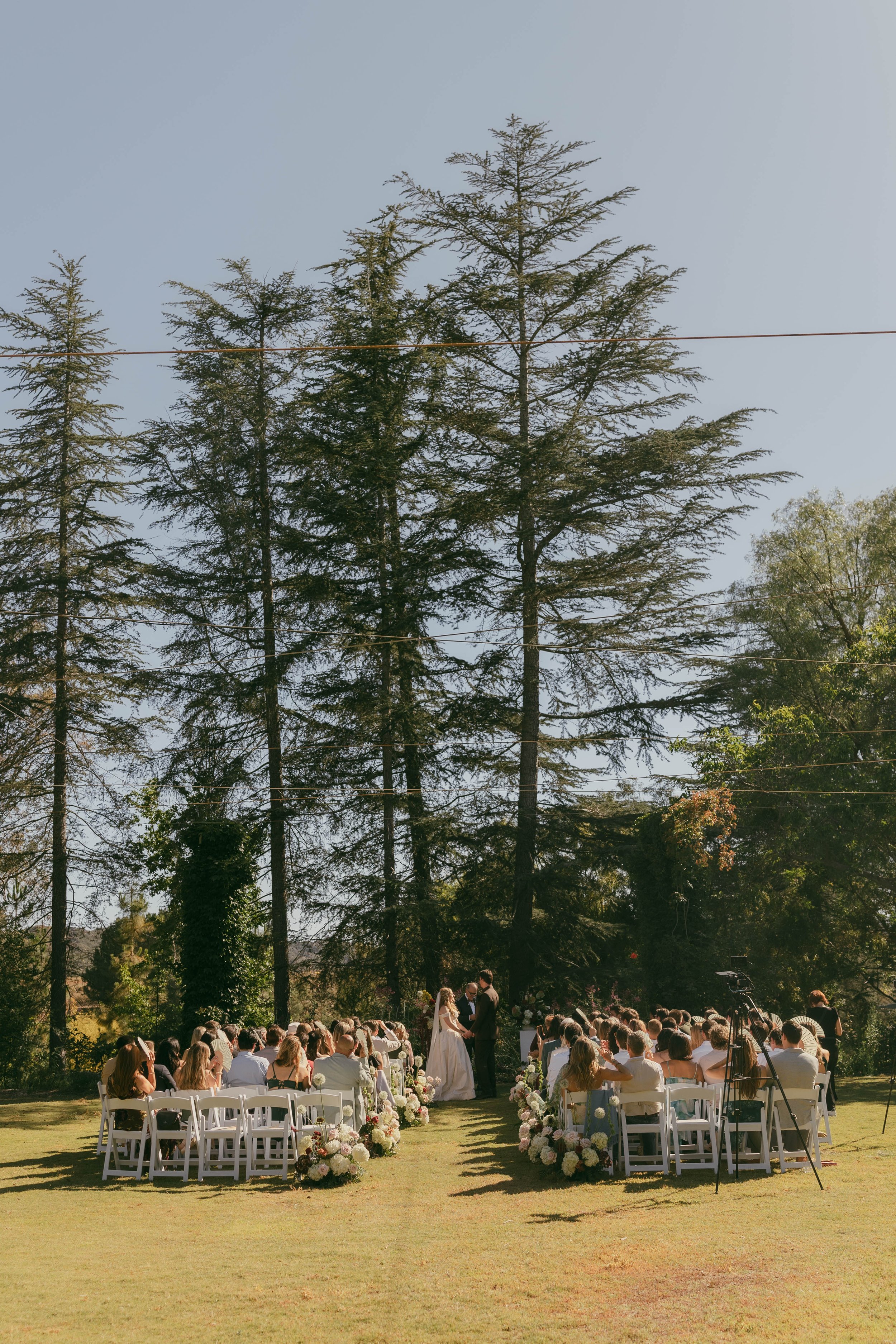 An outdoor wedding ceremony with a bride and groom standing before a large gathering of guests seated on white chairs, surrounded by floral arrangements, under tall trees on a sunny day.