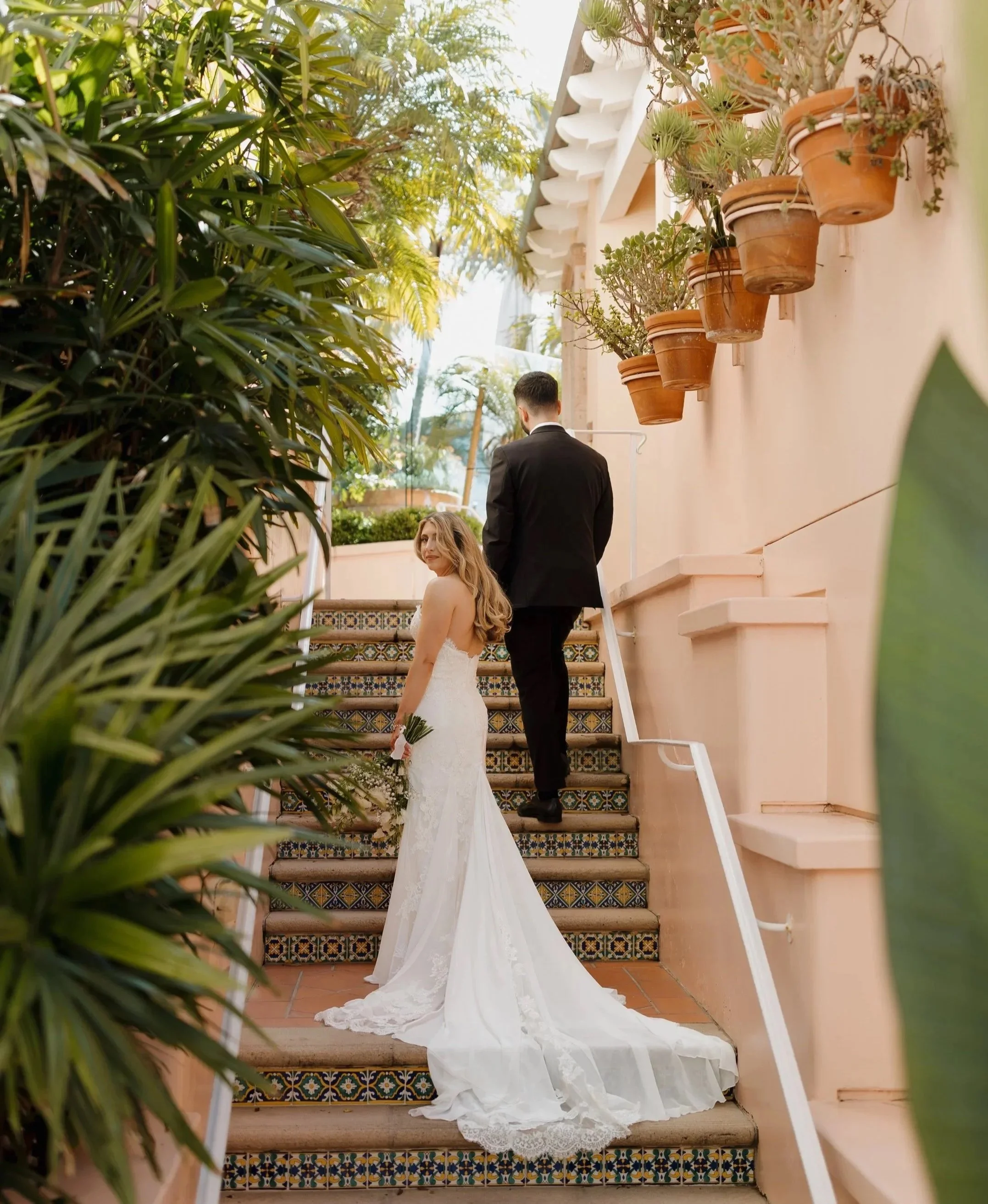 A bride in a white wedding gown with a long train, holding a bouquet, standing on decorative tiled stairs next to a groom dressed in a black suit, outdoors with plants and a peach-colored building.