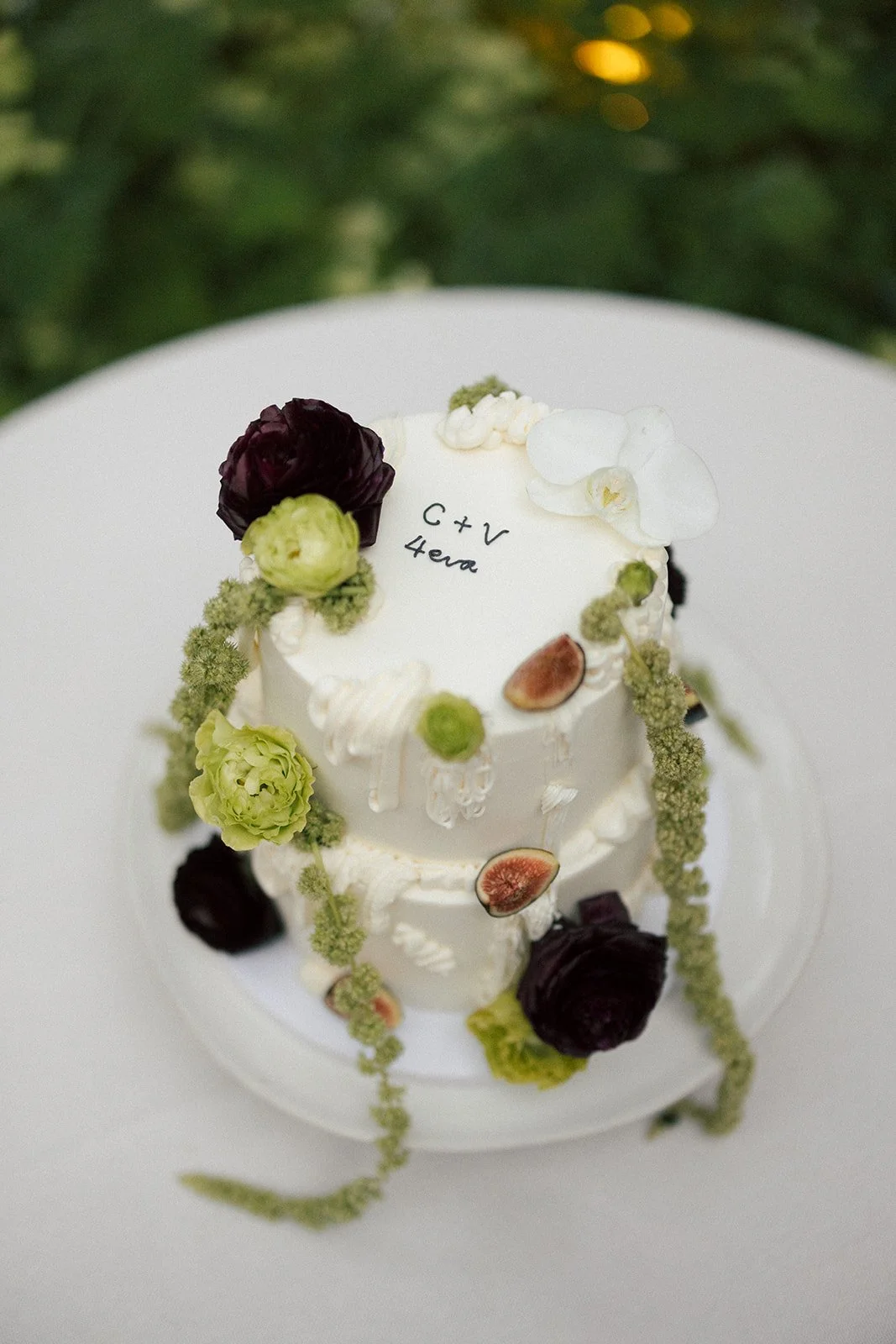 A white, two-tiered wedding cake decorated with dark purple and light green flowers, figs, and green hanging elements, with handwritten black text on the top tier that reads 'C + V 4eva'.