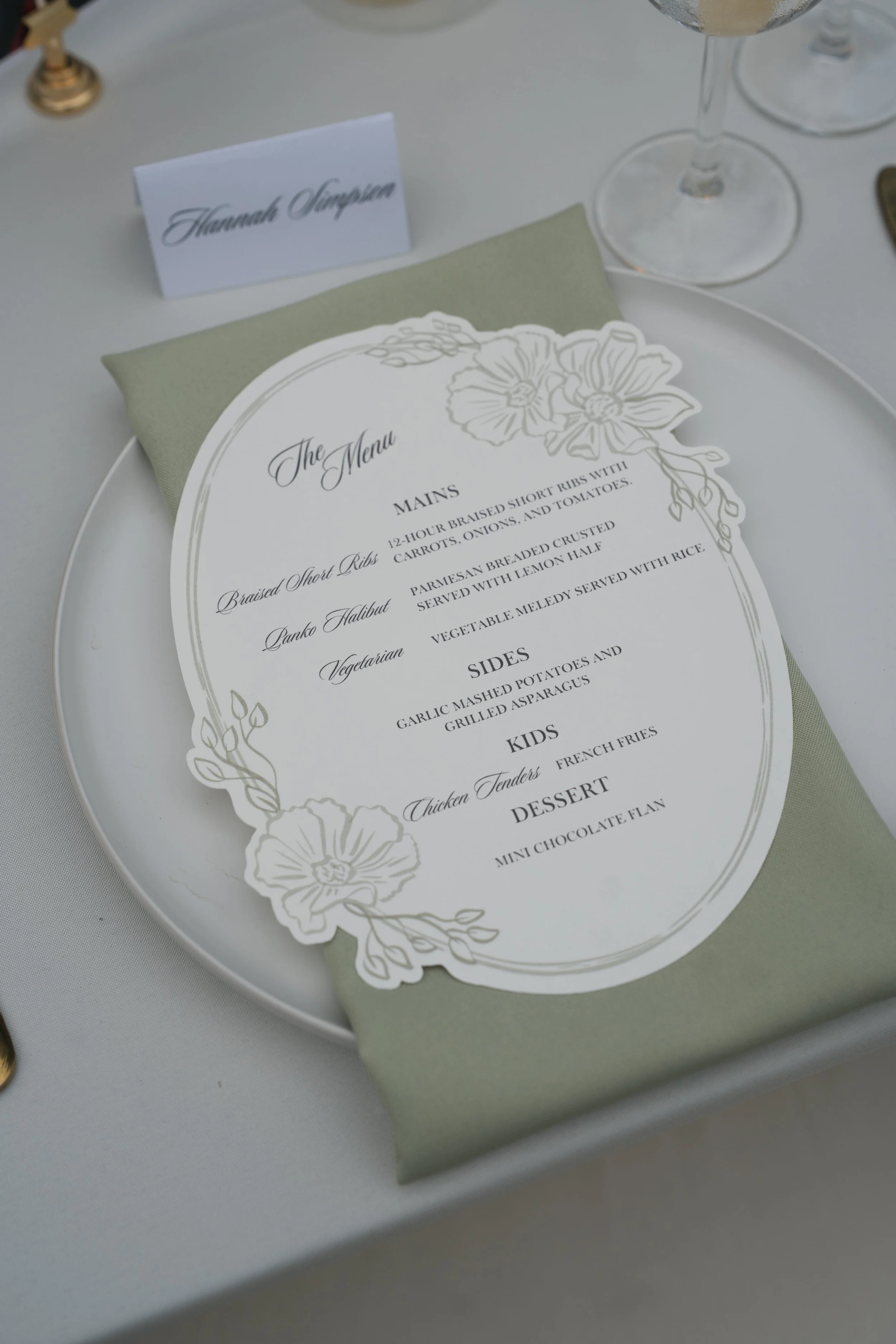 Wedding reception table with a menu card, a place card with the name Hannah Simpson, and glasses of wine or champagne.