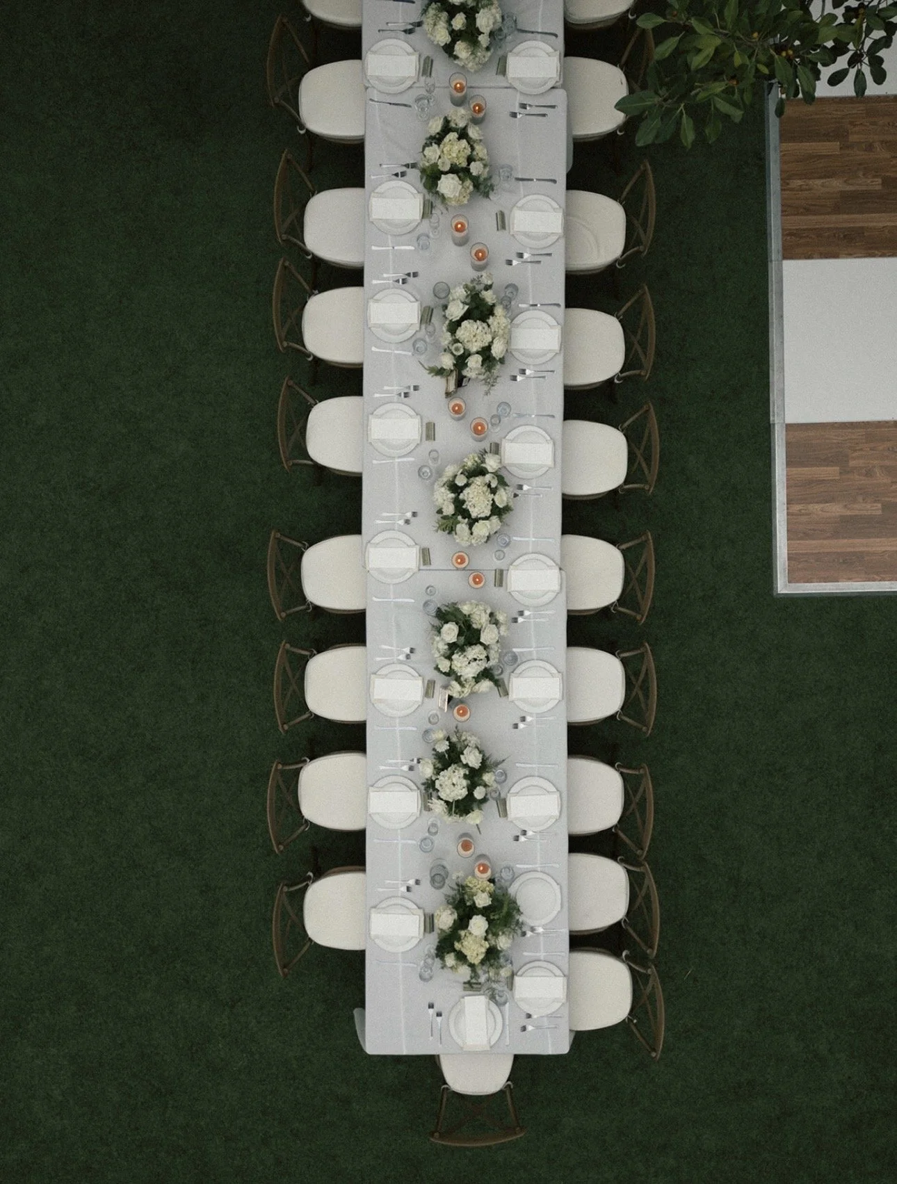 Overhead view of a long banquet table set for an event, decorated with white floral centerpieces and candles, on a green grass floor.
