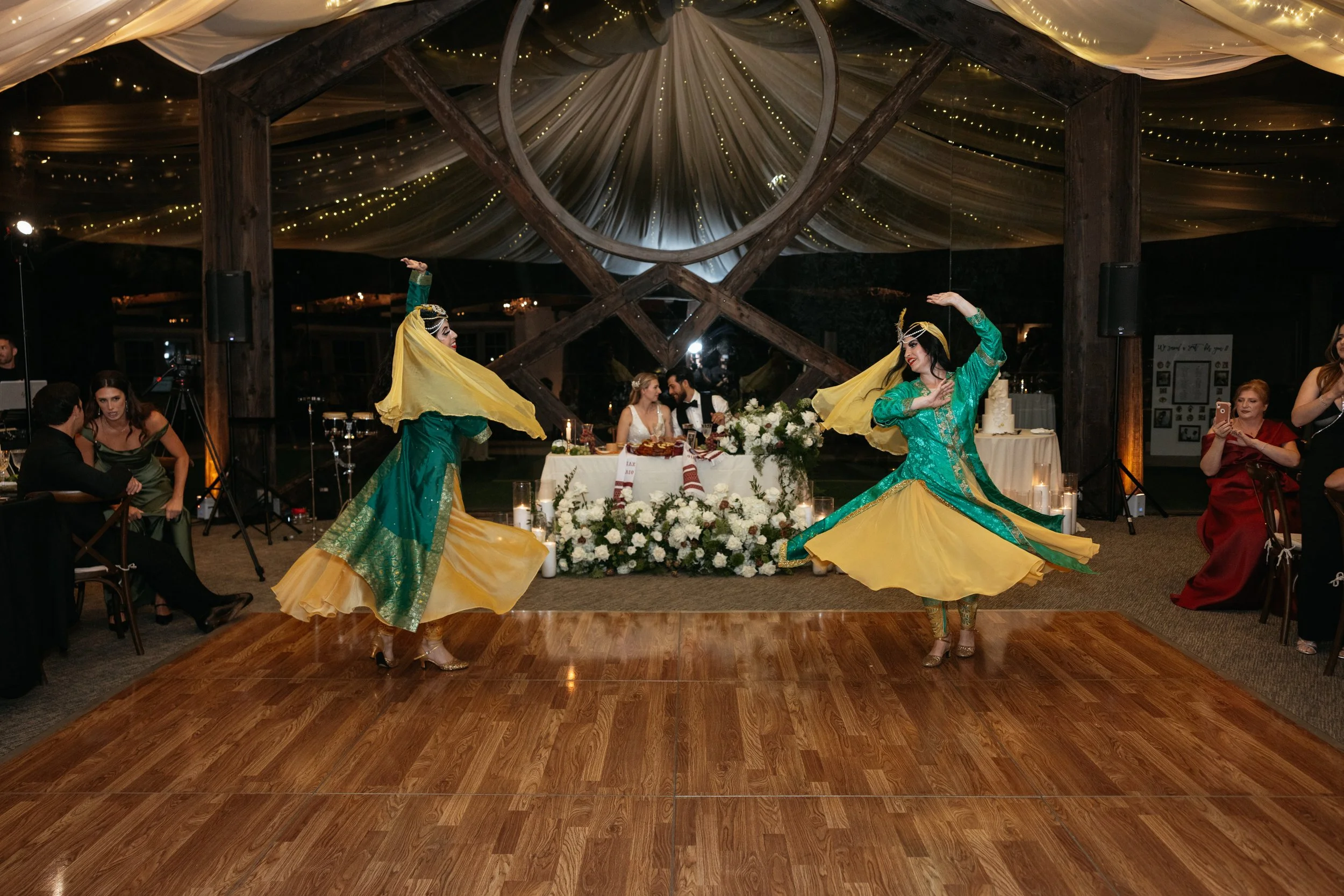 Two women in traditional costumes dancing on a wooden dance floor at a wedding reception. A bride and groom are sitting at a table decorated with white flowers and candles. Guests are seated around, some taking photos or watching the performance. The
