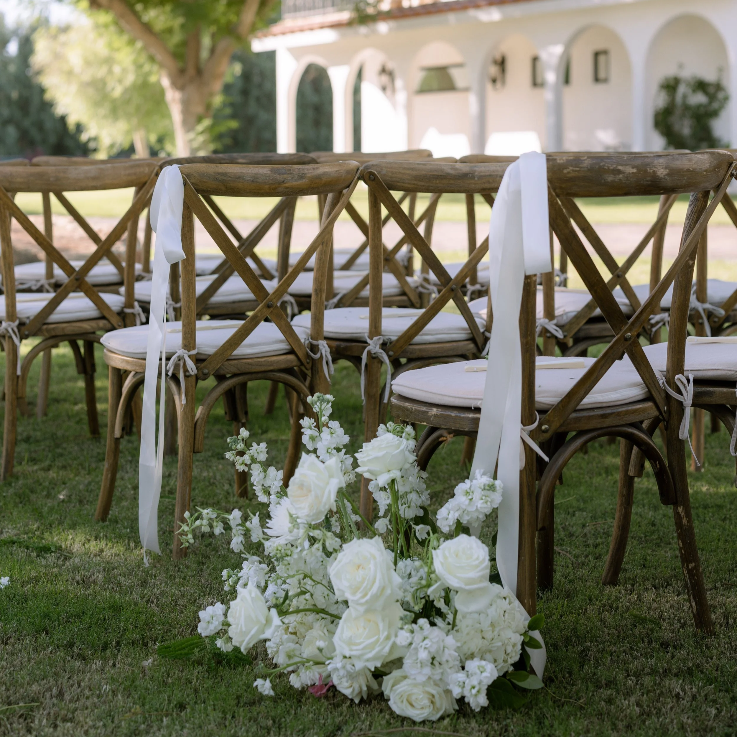 Outdoor wedding setup with wooden chairs decorated with white ribbons and a floral arrangement of white roses and other white flowers on the grass.