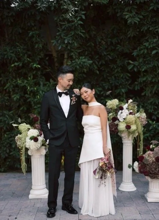 A bride and groom standing together at their wedding. The groom is dressed in a black tuxedo with a bow tie, and the bride is wearing a strapless white wedding gown holding a bouquet of flowers. They are outside, with a backdrop of greenery and flowers in white and deep red on pedestal stands.