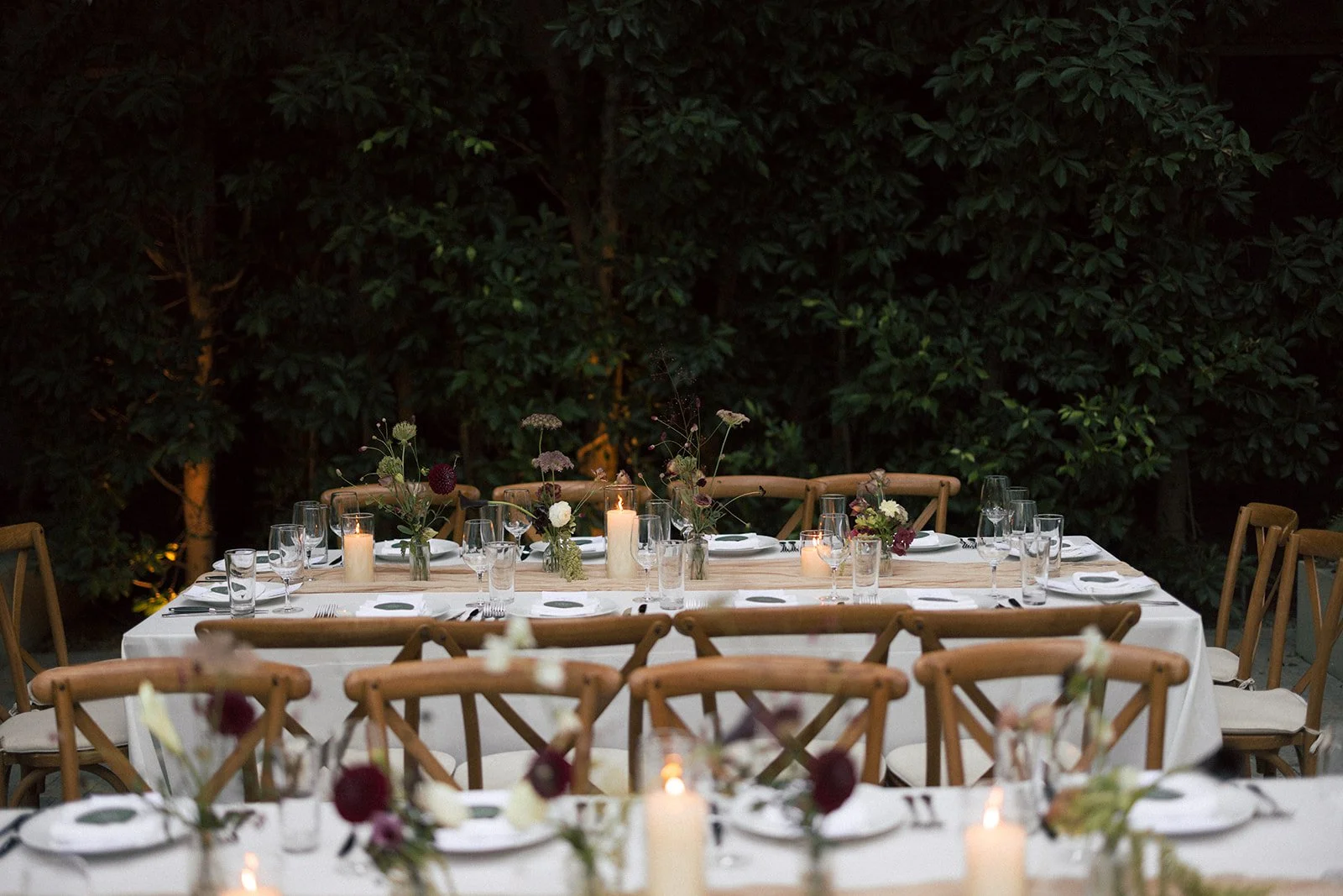 A long dining table set for an event with candles, floral centerpieces, glasses, plates, and silverware, outdoors against a backdrop of lush green trees.