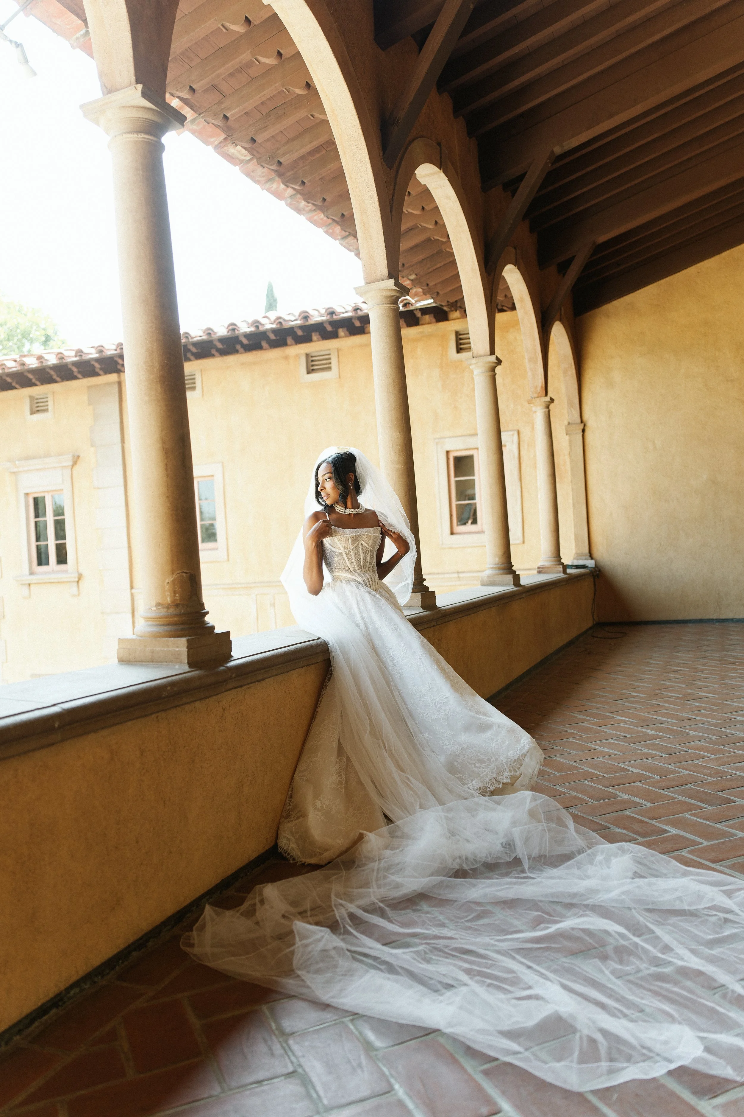 A bride in a white wedding gown with a long train, sitting on a ledge in a covered walkway with stone columns and a tiled roof, holding her veil and looking to the side.