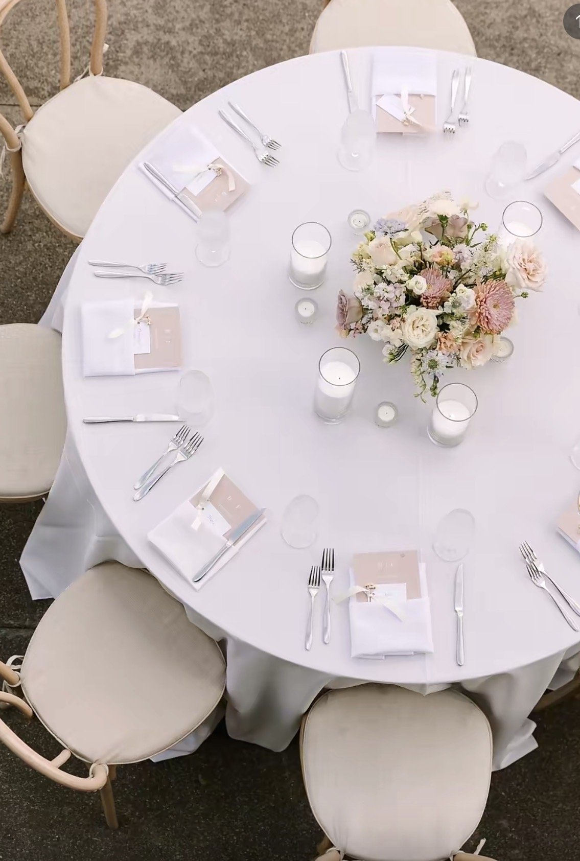 A round banquet table set for a formal event with six place settings, a floral centerpiece, candles, and wrapped menus, surrounded by six beige cushioned chairs.