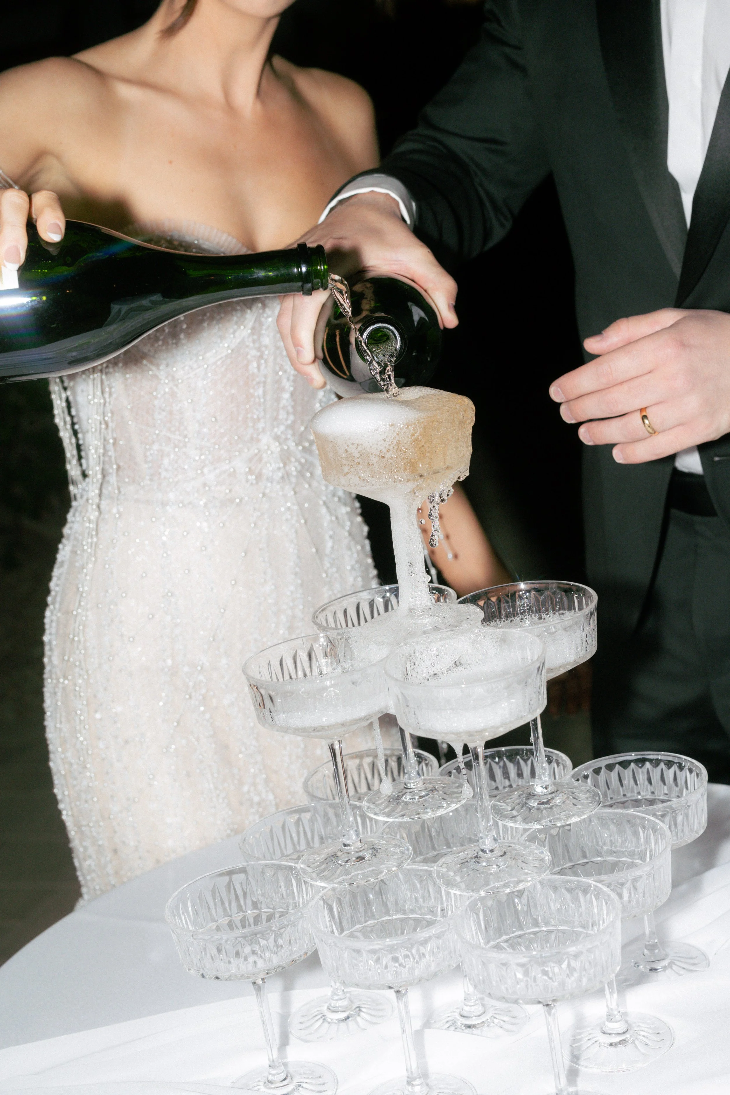A bride and groom pouring champagne into a tower of glasses at their wedding reception.
