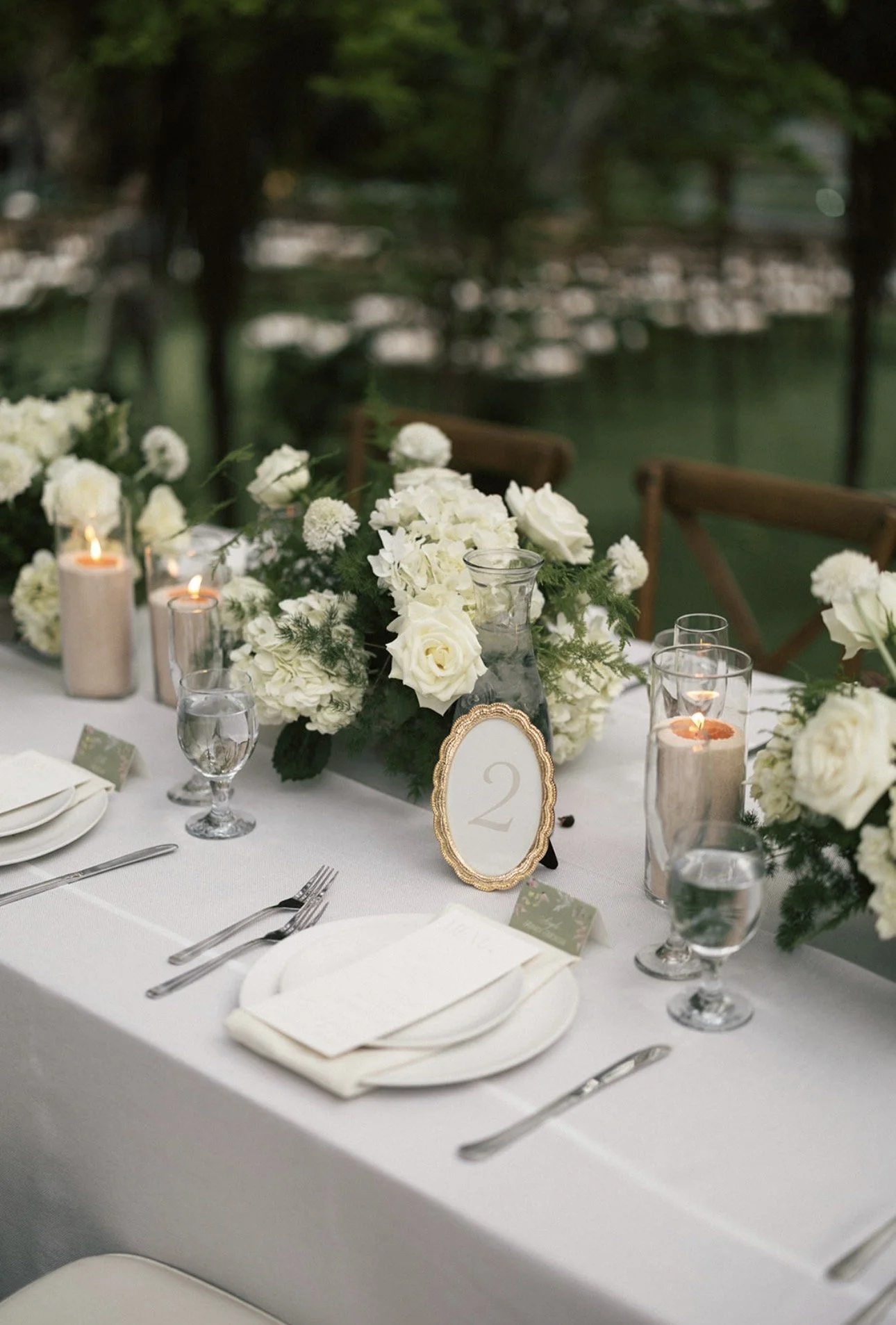 Elegant dining table setup with white floral centerpiece, candles, glassware, and place settings, outdoors with water and trees in the background.