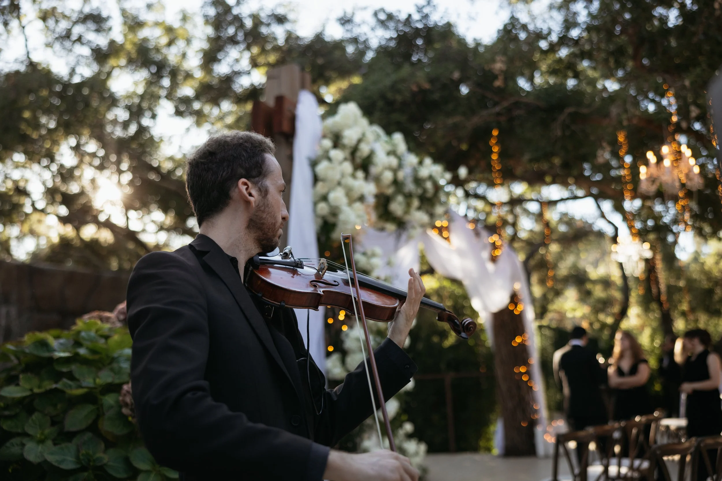 A man in a black suit playing the violin at an outdoor wedding ceremony, with a decorated arch, white flowers, trees, and warm string lights in the background.