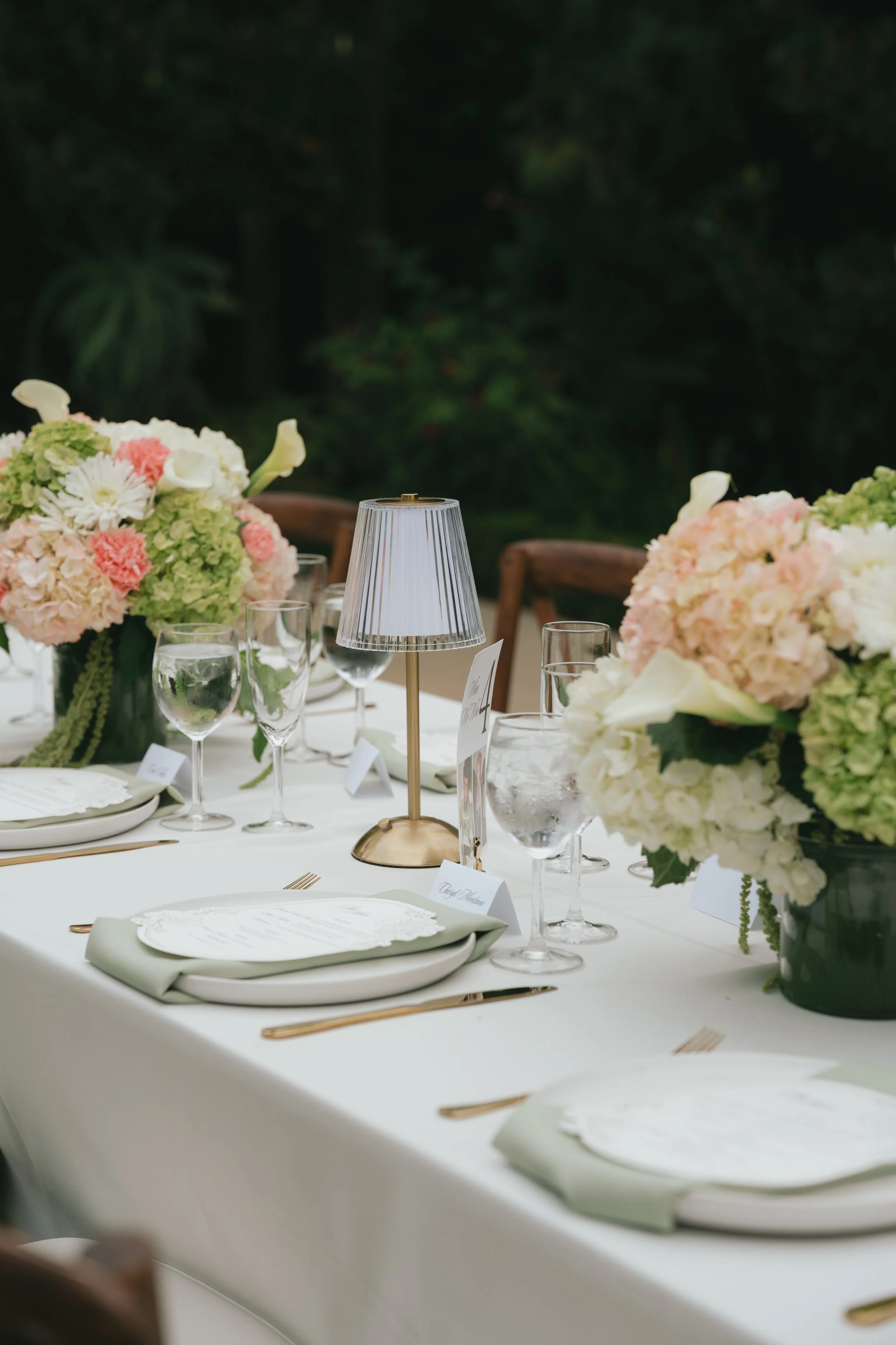 Elegant wedding reception table decorated with pink and white floral centerpieces, gold cutlery, white plates with folded napkins, and a small lampshade centerpiece, set outdoors with a lush green background.