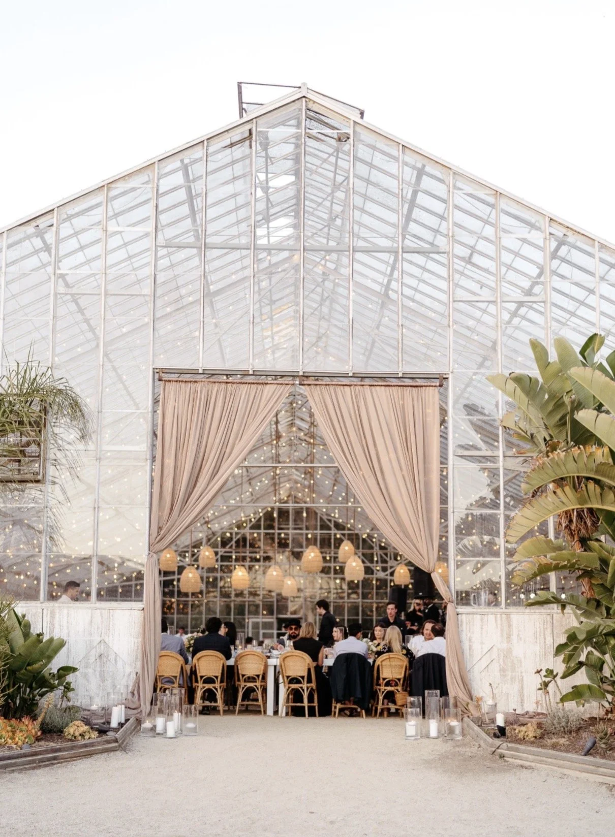 Glass greenhouse wedding reception with beige curtains, string lights, and guests seated at tables.