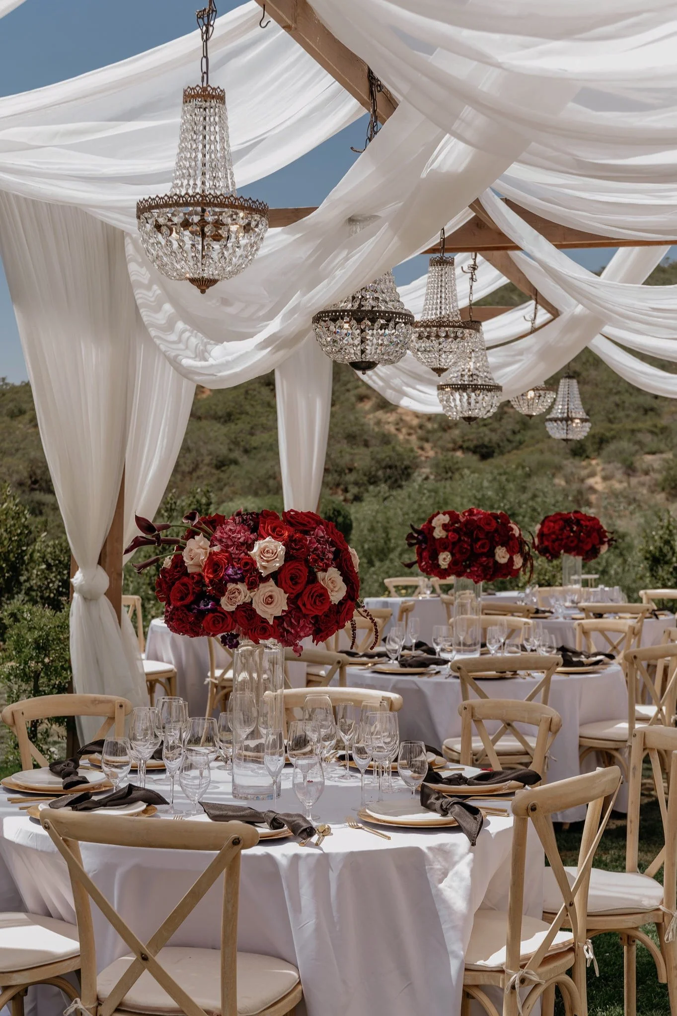 A decorated outdoor event space with white draped fabric and chandeliers hanging from a wooden frame. There are tables with white tablecloths, glassware, and floral centerpieces with red, white, and purple flowers.